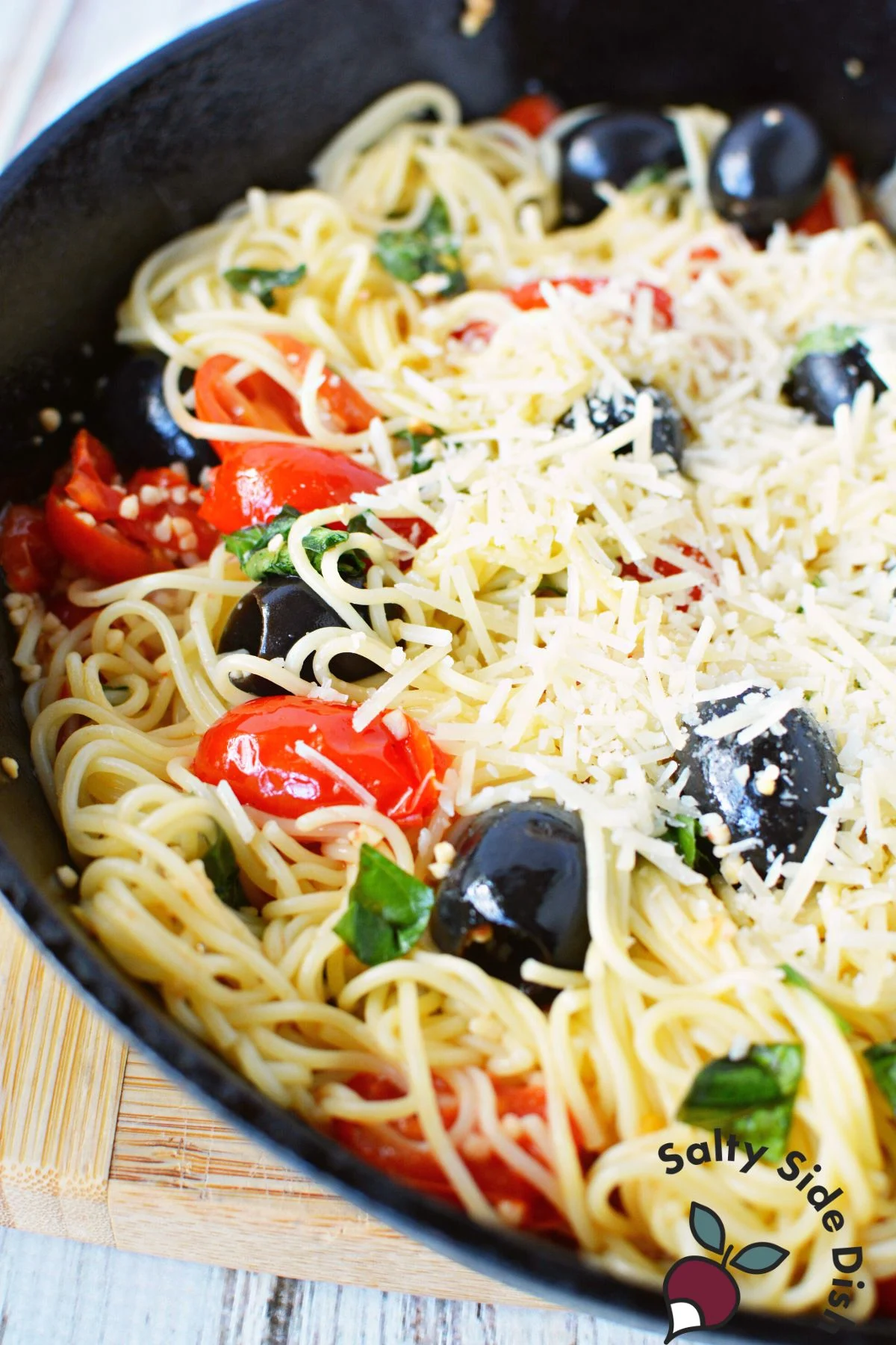 angel hair pasta with tomatoes, full olives, and parmesan cheese in a cast iron pan.