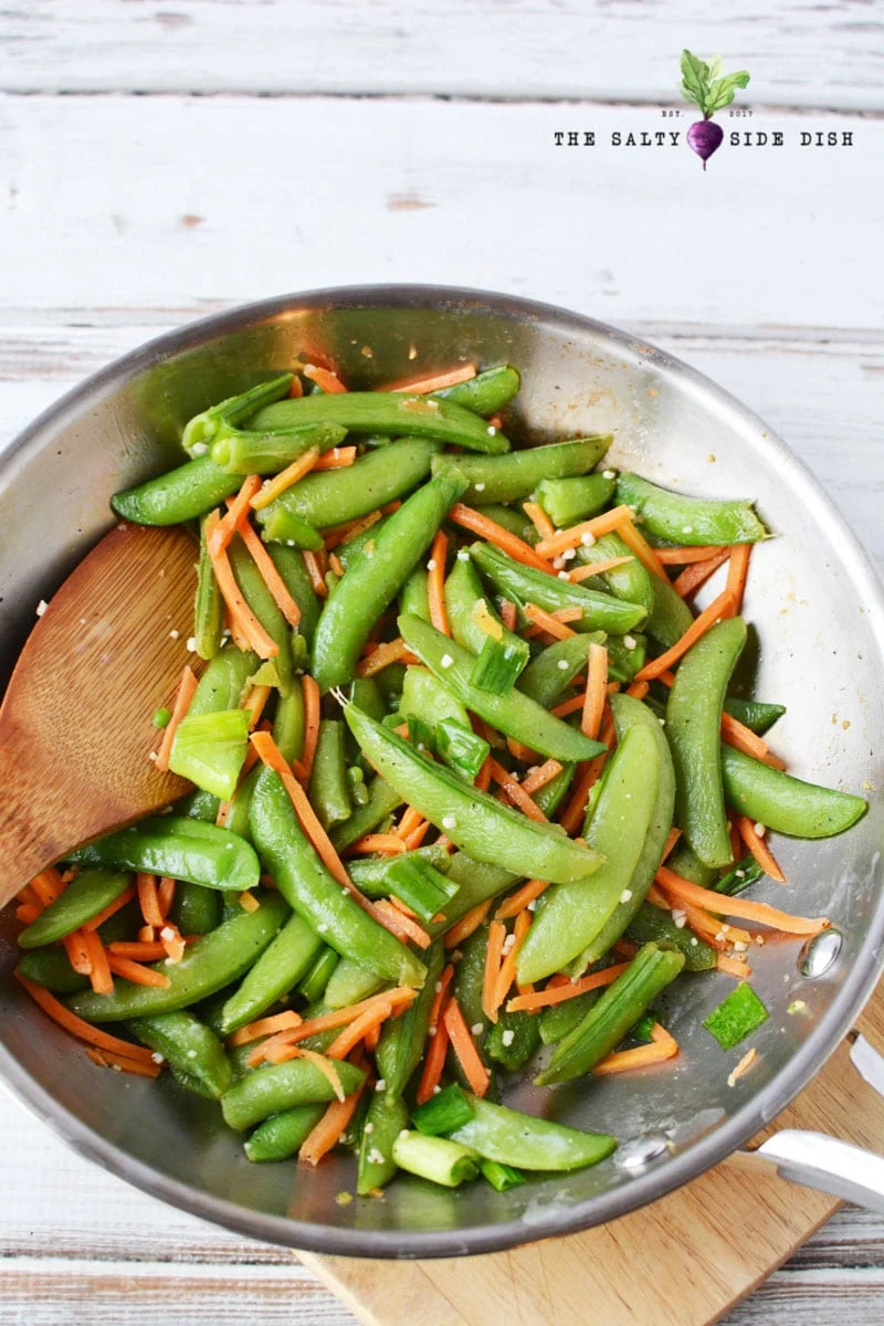 sauteed sugar snap peas in skillet with garlic and matchstick carrots being stirred by a wooden spoon