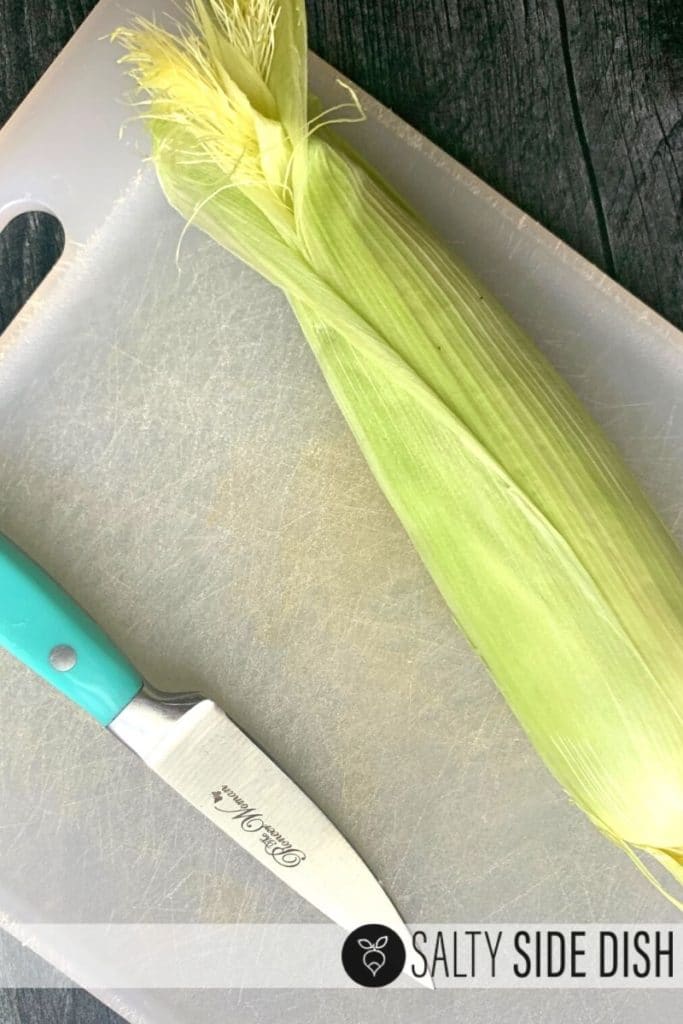 An ear of corn on a cutting board with a knife.