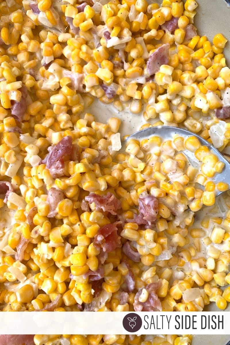 Creamy Corn and Crispy Bacon Smothered in Shaved Parmesan in a pan with a spoon to scoop out this side dish