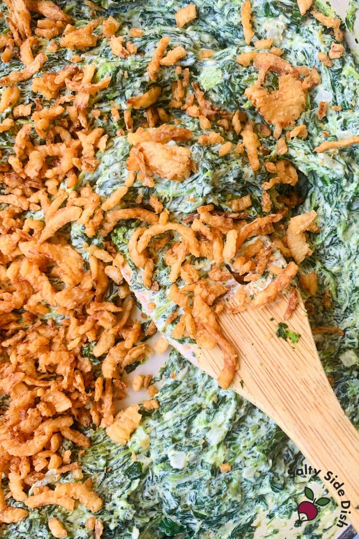 pan with wooden spoon stirring creamed spinach with a lot of French's crunchy fried onions on top