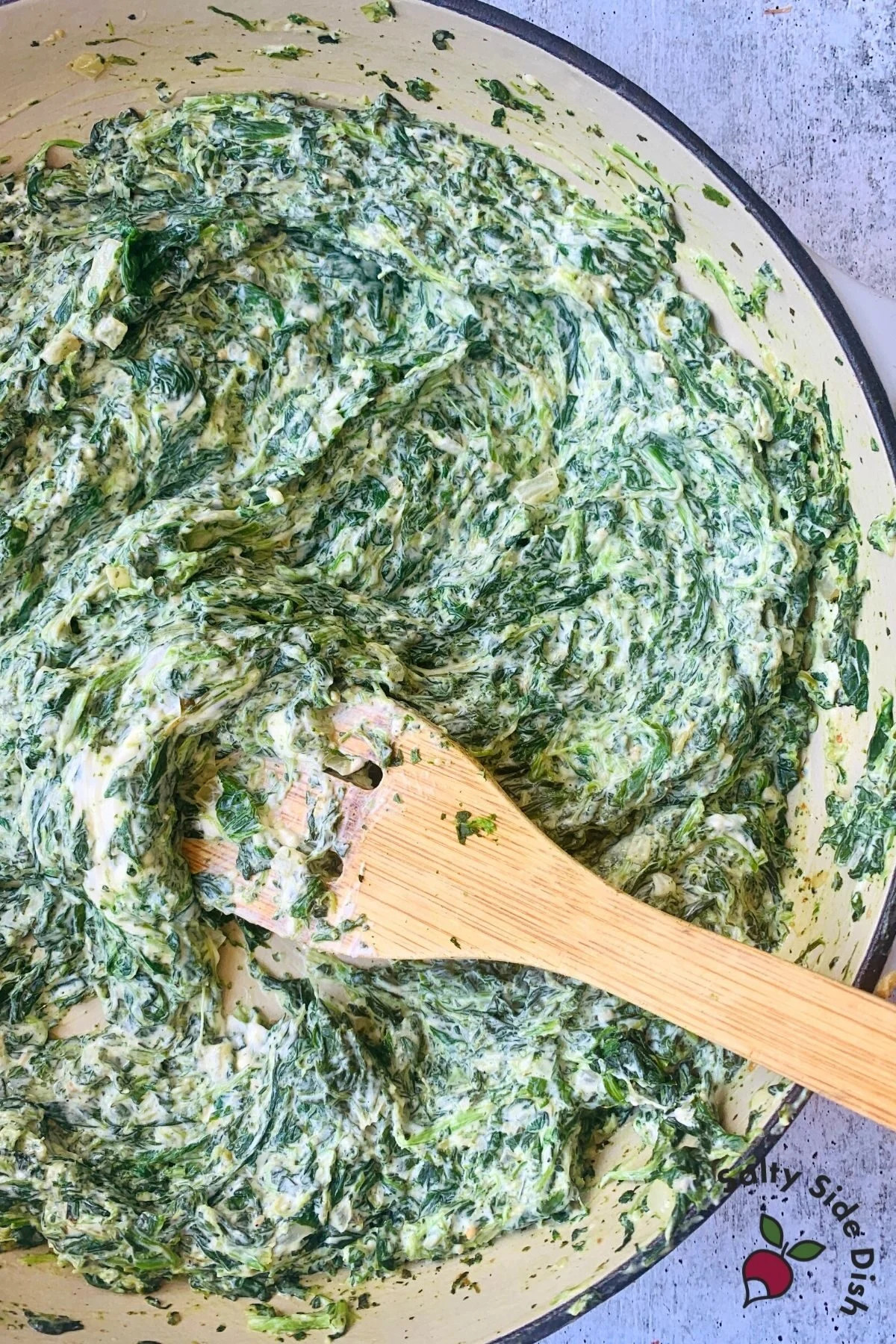 creamed spinach in a cast iron pan being stirred with a wooden spoon