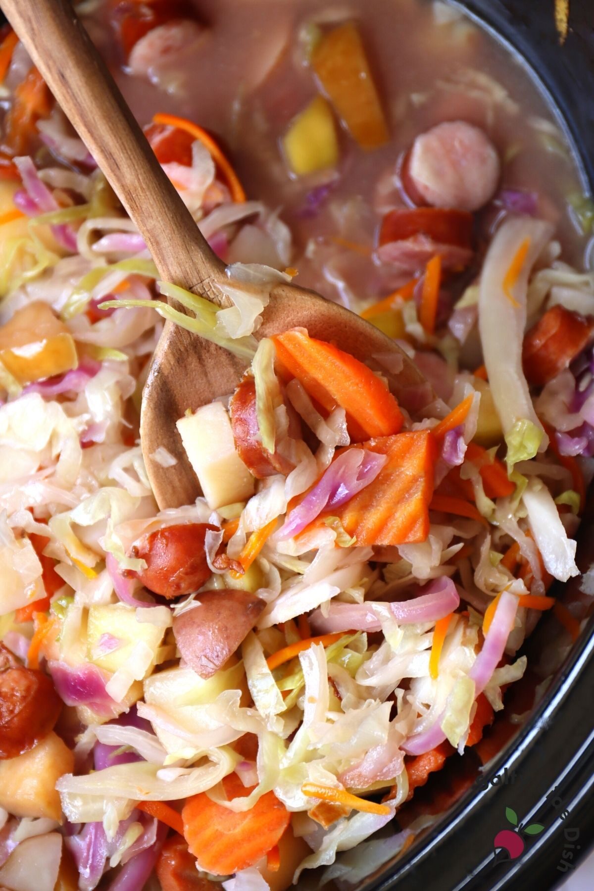 large slow cooker with sausage and cabbage stew on a wooden spoon