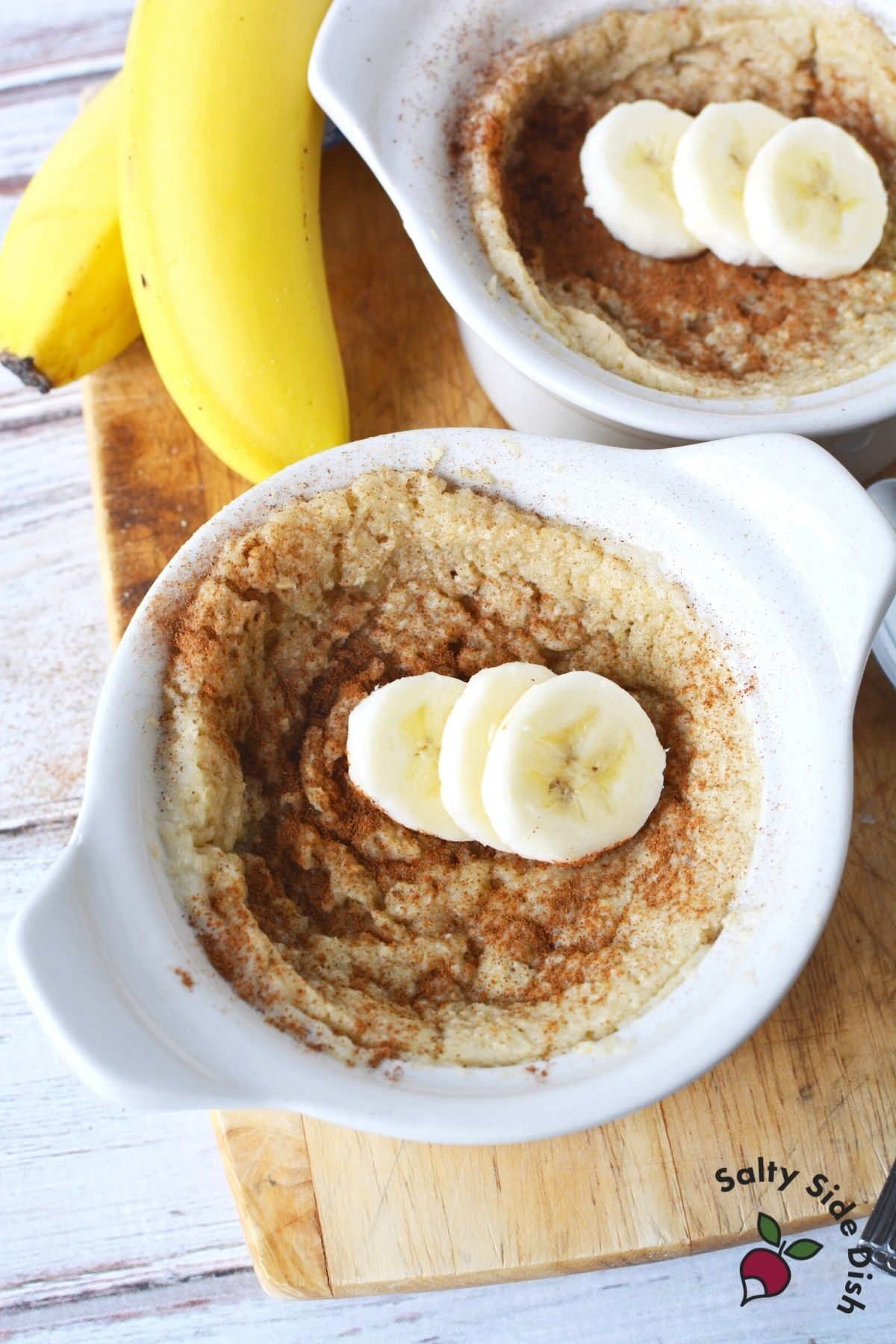 two white ramekins with baked oats, banana slices on top and banana in background left.