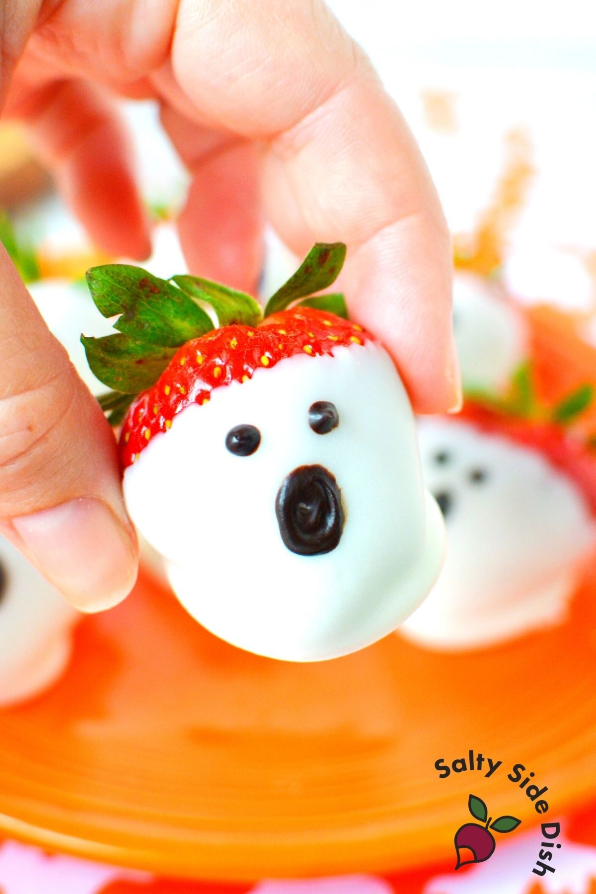 fingers holding a chocolate dipped strawberry that looks like a ghost with an orange plate in background.
