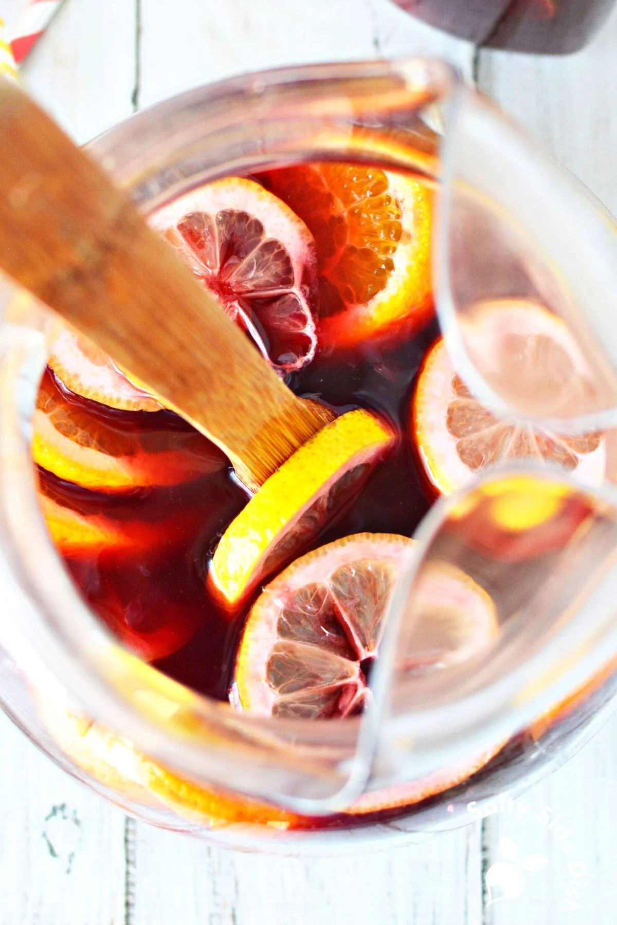 fresh merlot sangria in a pitcher with fresh fruit slices on top being stirred by a wooden spoon.