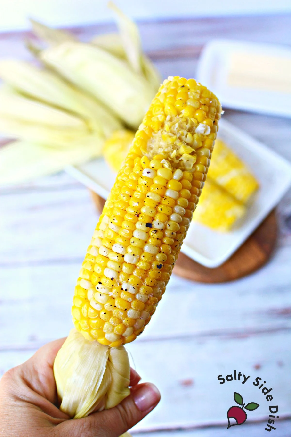 A hand holding an ear of corn on the cob by the husk. The corn has a bite taken out of it.