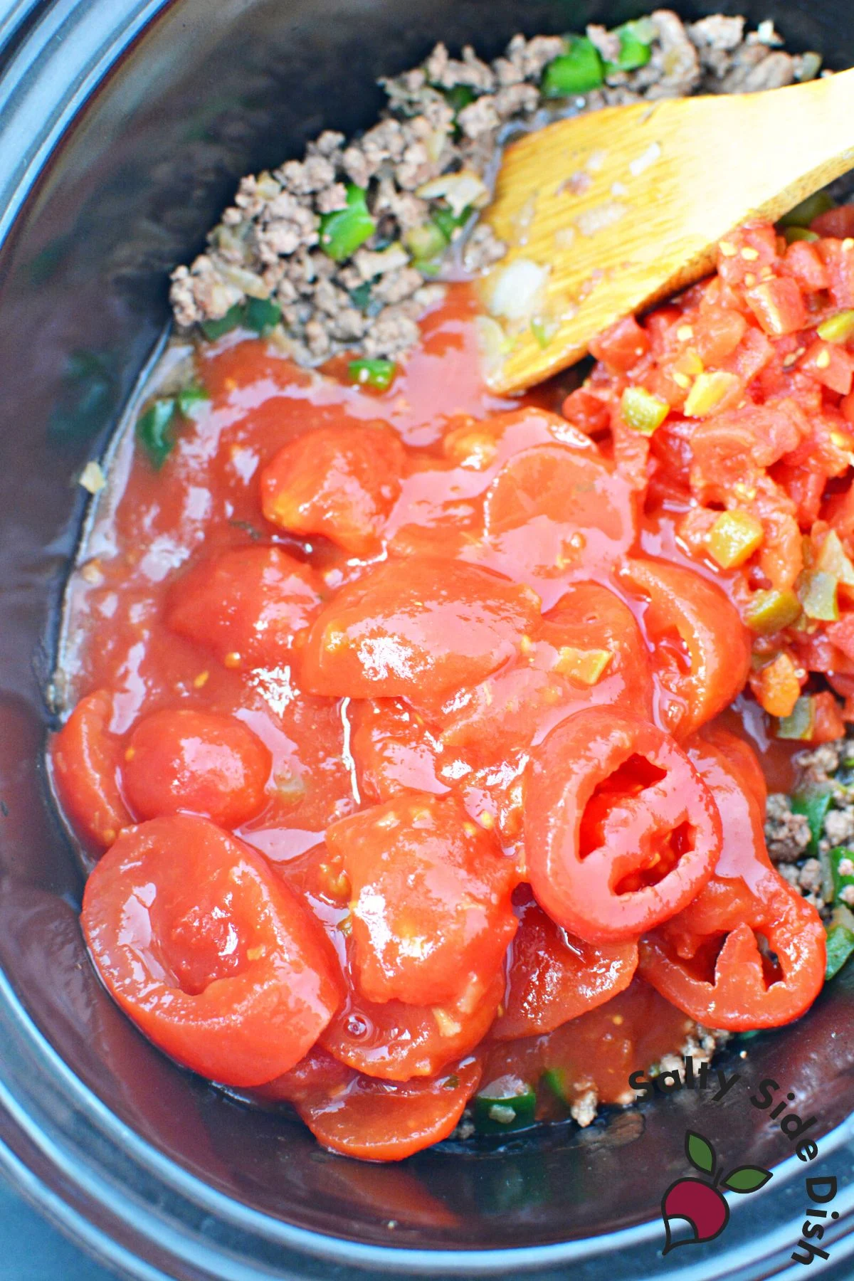slow cooker with tomatoes and beef being stirred by a spoon.