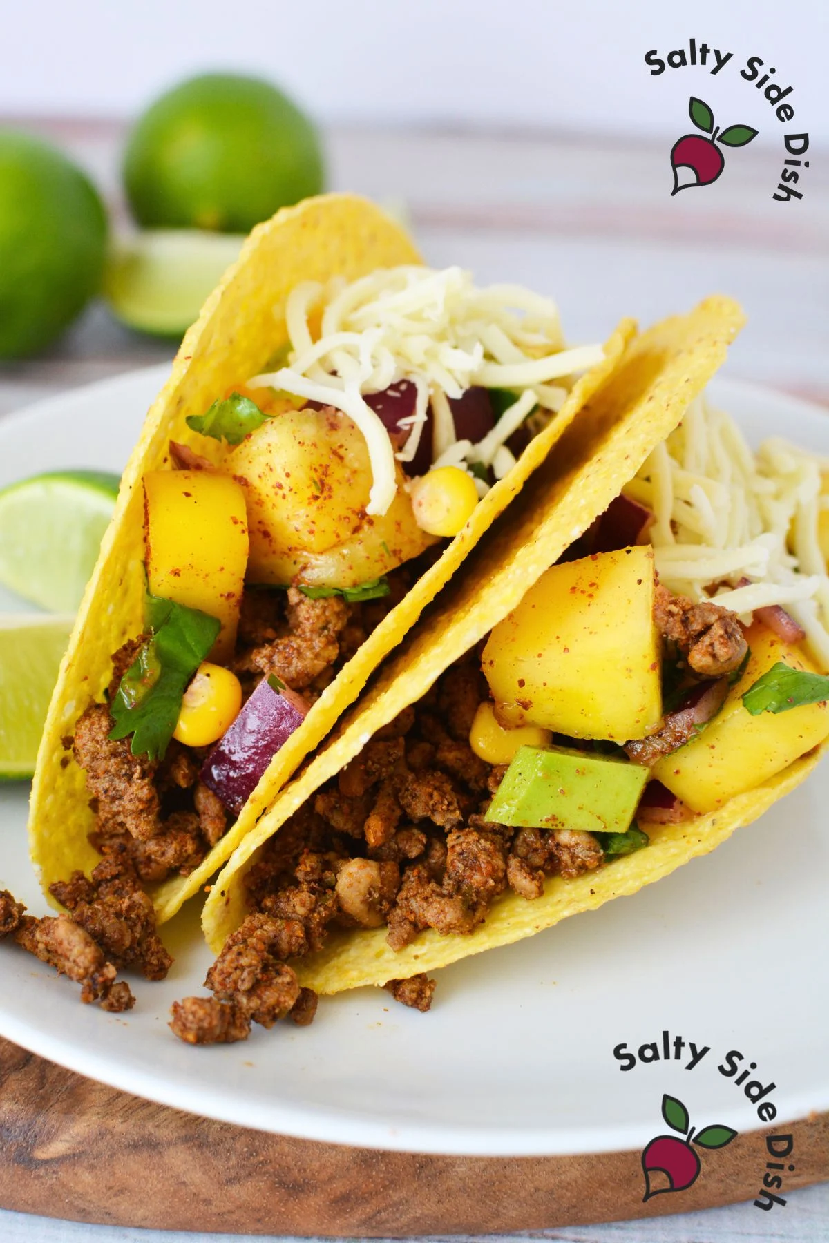A fresh and colorful Pineapple Mango Fruit Salad used as a topping on tacos, featuring diced pineapple, mango, corn, red onion, jalapeno, avocado, and cilantro, garnished with lime wedges on a taco shell.
