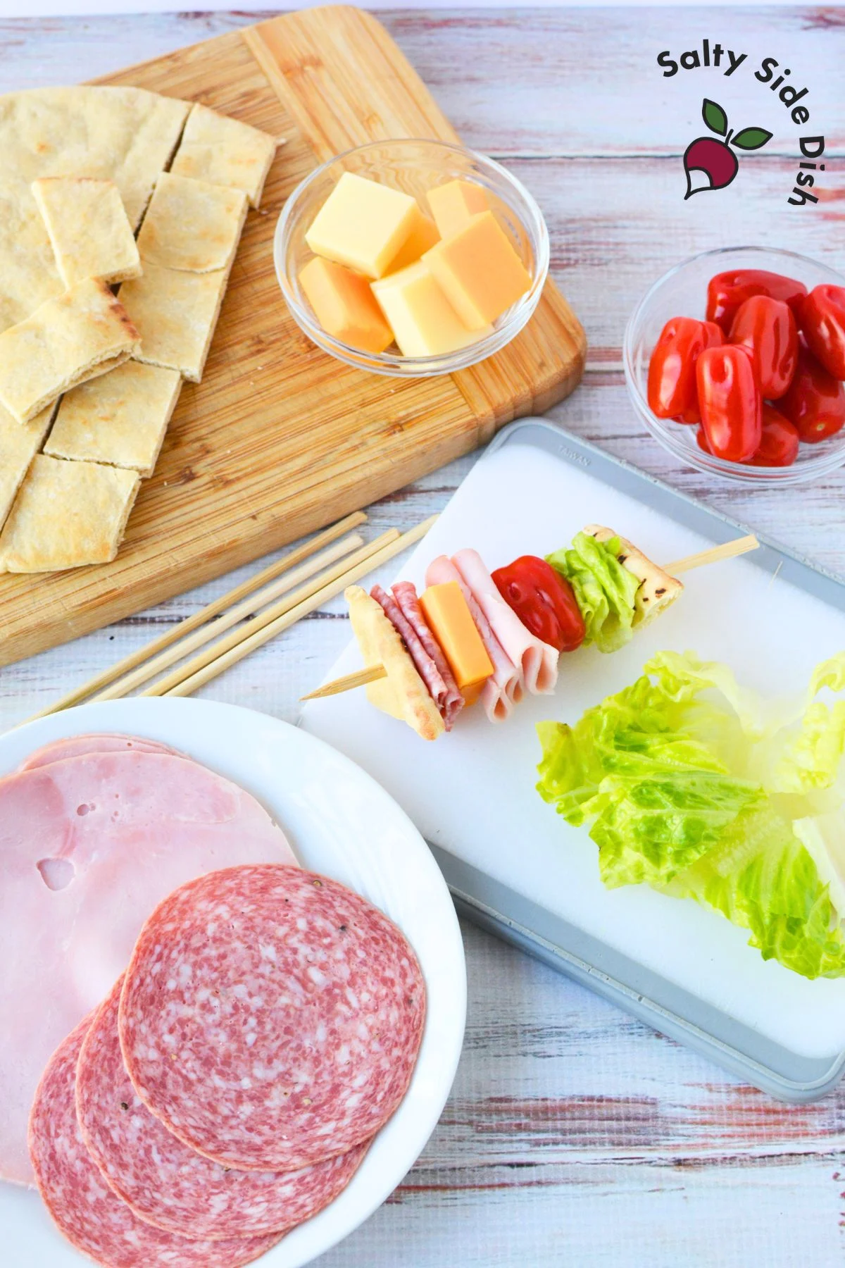 A variety of ingredients laid out for sandwich on a stick, including sliced pita bread, chopped romaine lettuce, grape tomatoes, sliced salami, sliced ham, and cheese cubes.