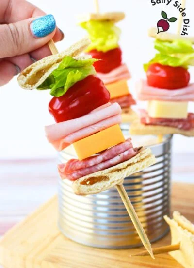Close-up view of ingredients for sandwich on a stick, including sliced pita bread, chopped romaine lettuce, grape tomatoes, sliced salami, sliced ham, and cheese cubes, ready for assembly.