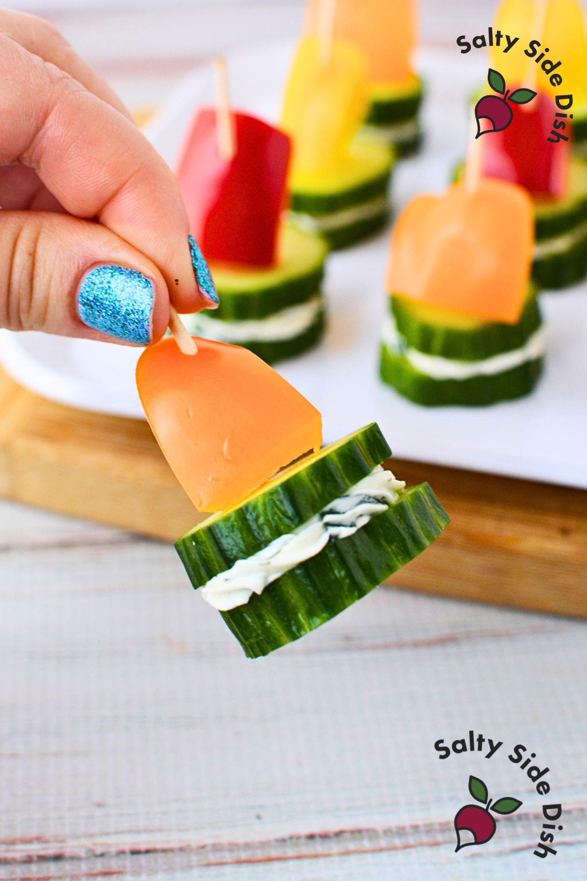 toothpick holding together cucumbers with topping of a bell peppers and cream cheese in center.