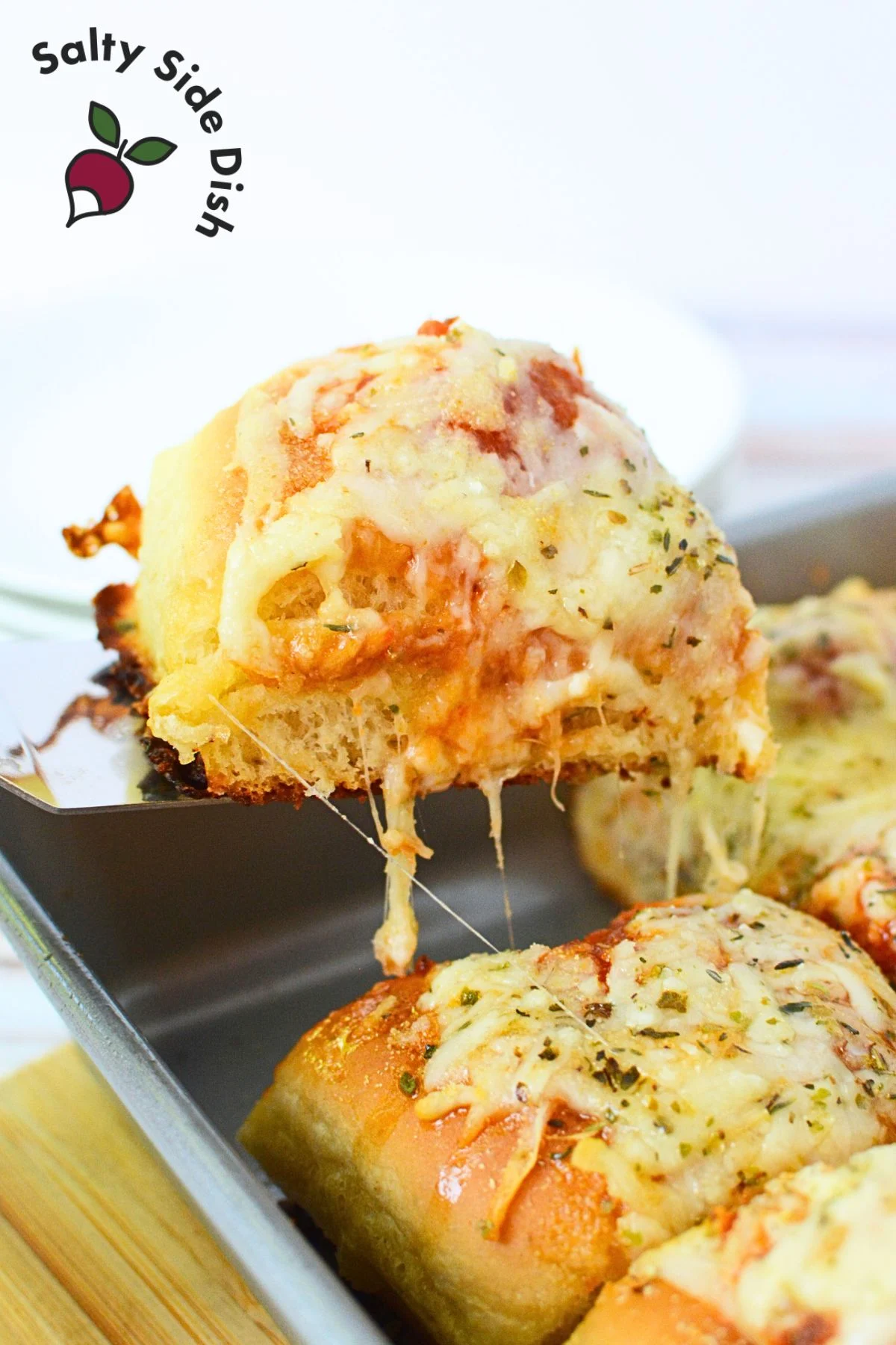 A meatball slider being lifted from the baking dish with a spatula, showing gooey cheese and a perfectly toasted Hawaiian roll.