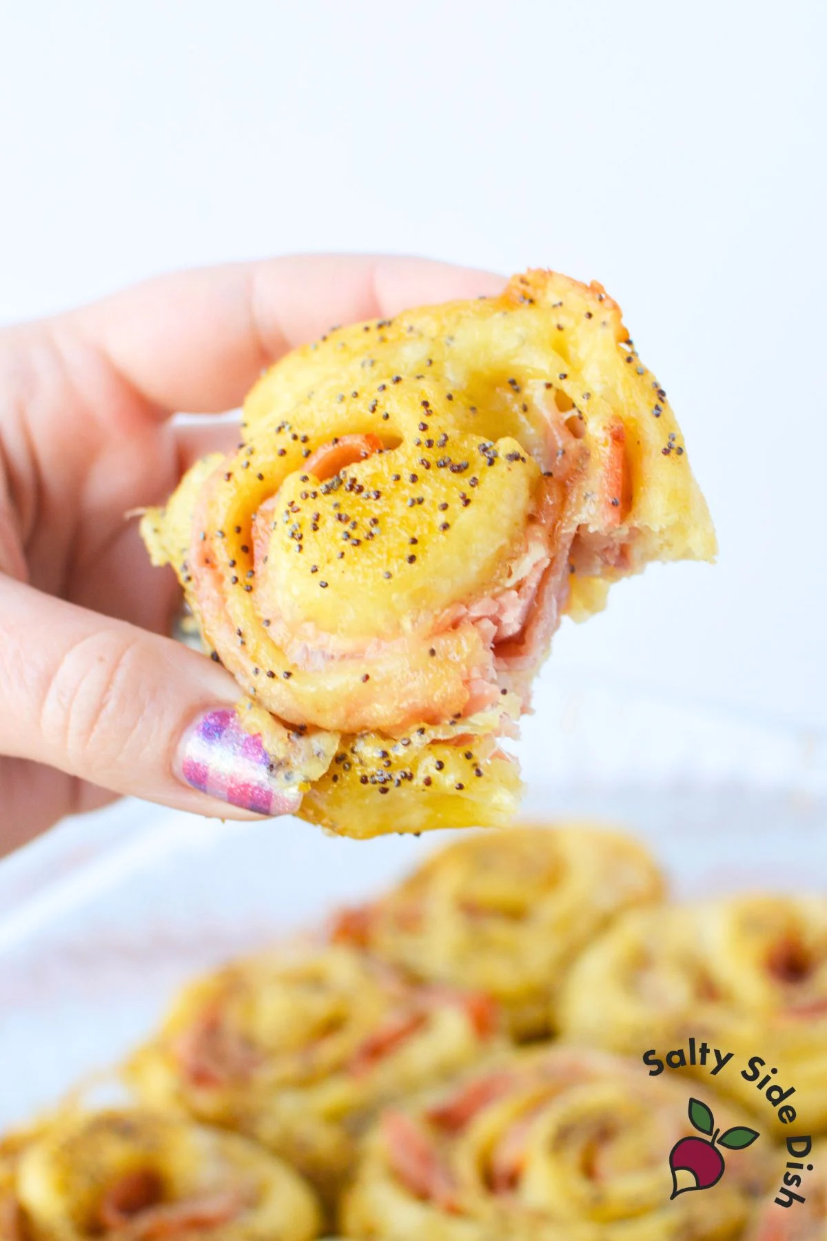 Hand holding a single ham and cheese pinwheel with visible layers of melted cheese, ham, and poppy seed glaze.