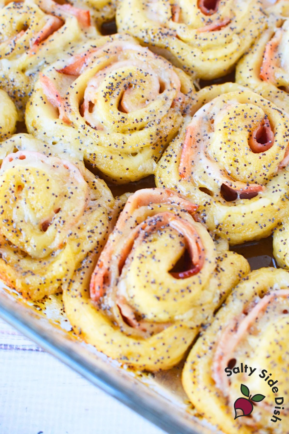 Full baking dish of golden-baked ham and Swiss pinwheels with buttery sauce at the bottom
