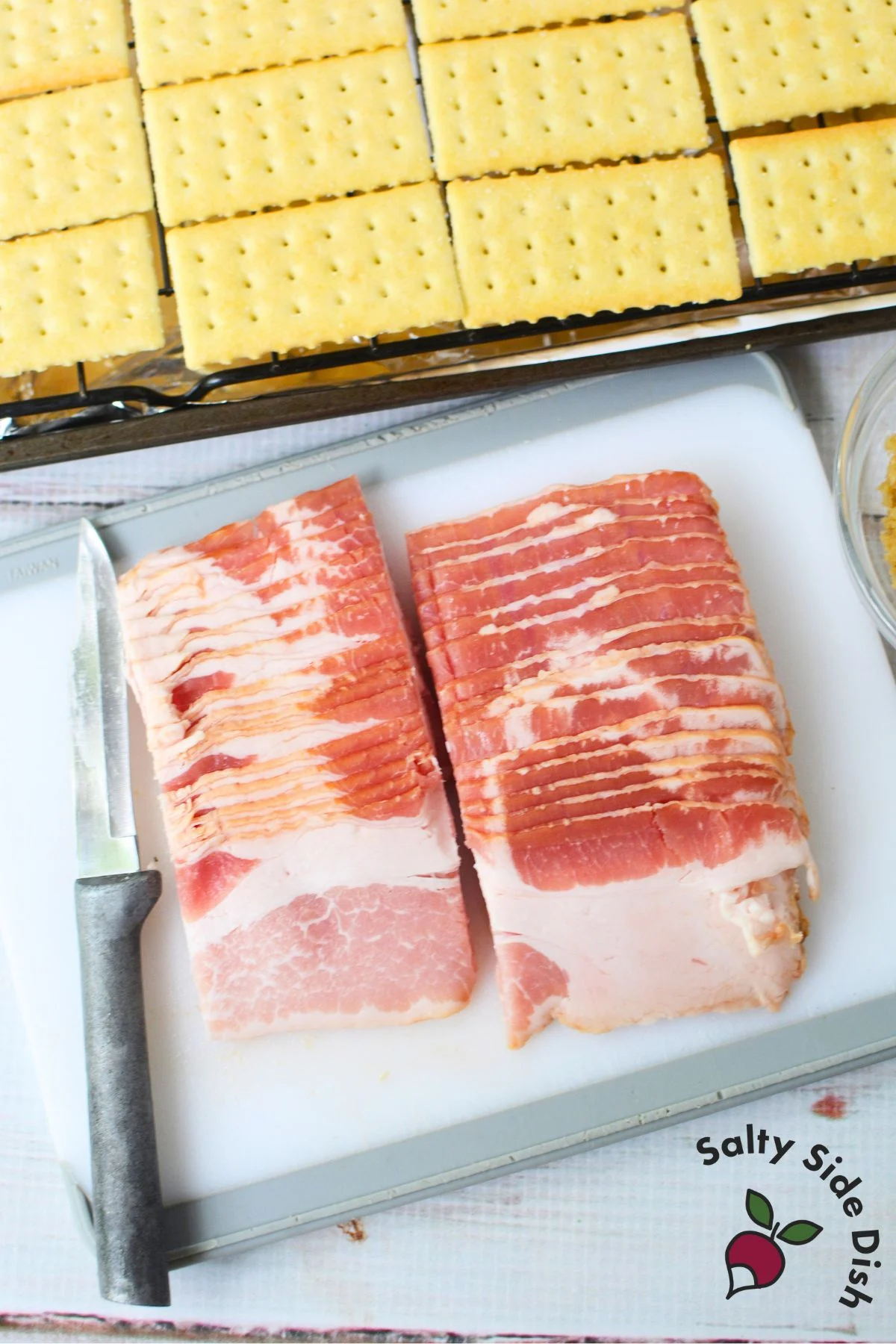Raw bacon being sliced into quarters on a cutting board next to club crackers for appetizer prep