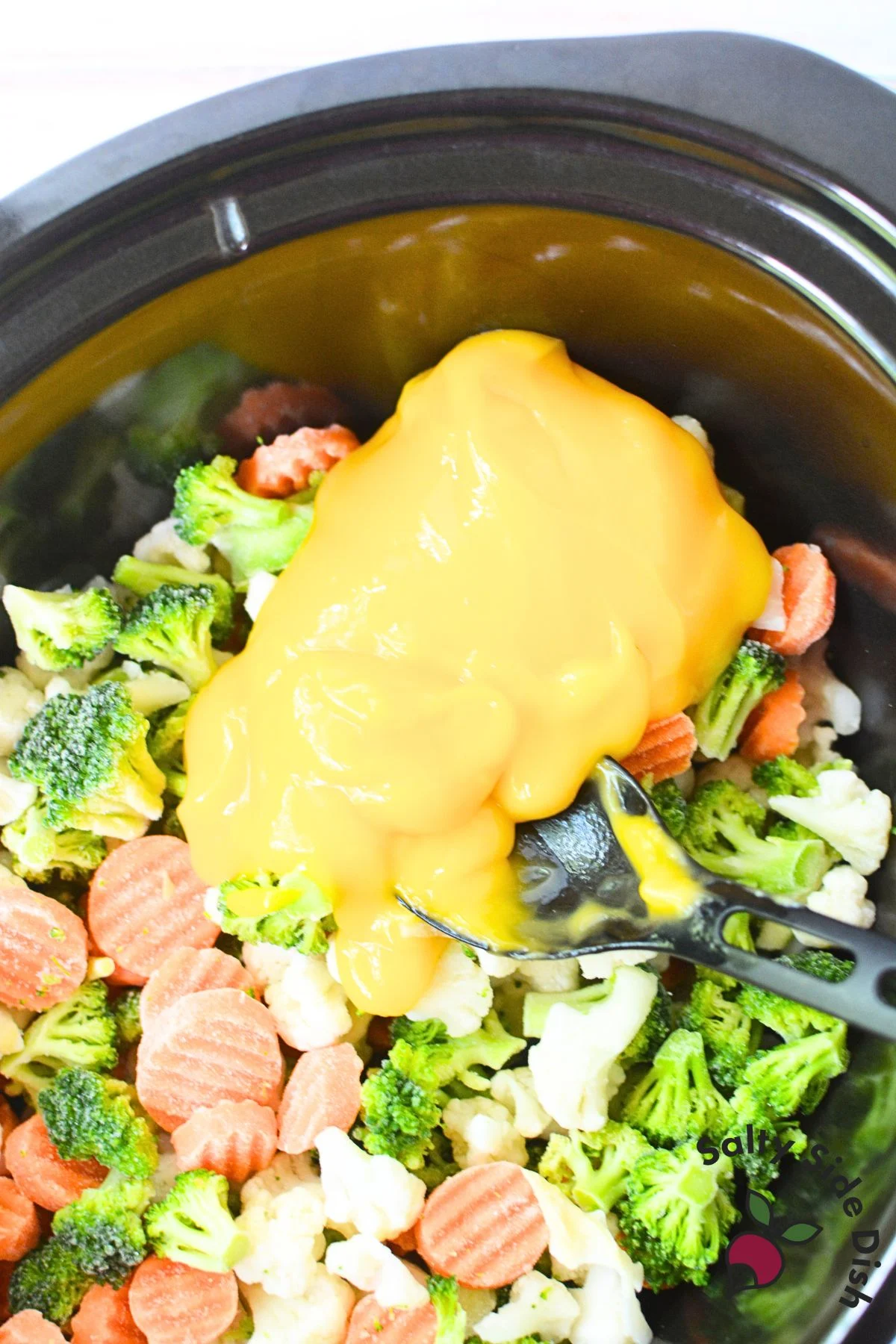 Cheddar cheese soup being added to frozen broccoli, carrots, and cauliflower in slow cooker