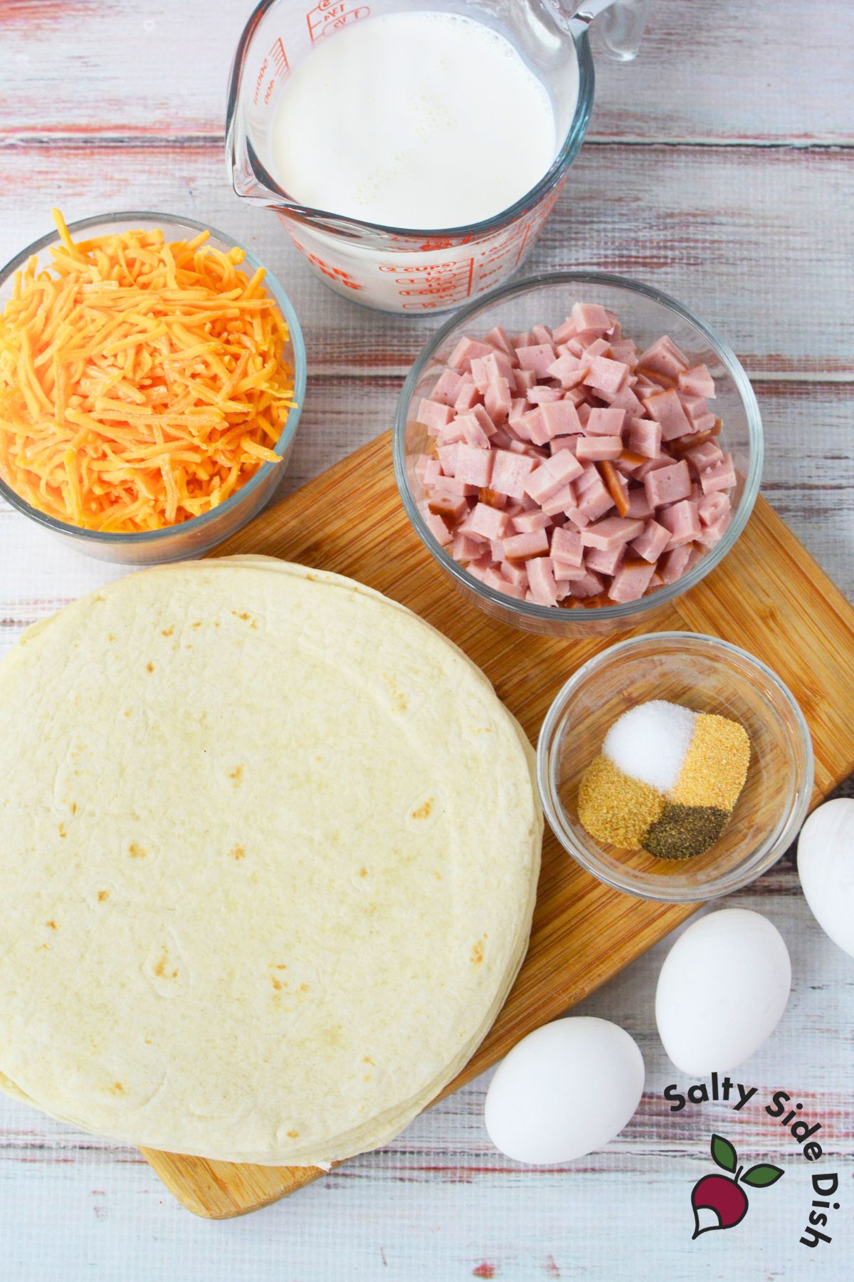 Ingredients for ham and egg breakfast tortillas with shredded cheddar, diced ham, eggs, half and half, and flour tortillas.