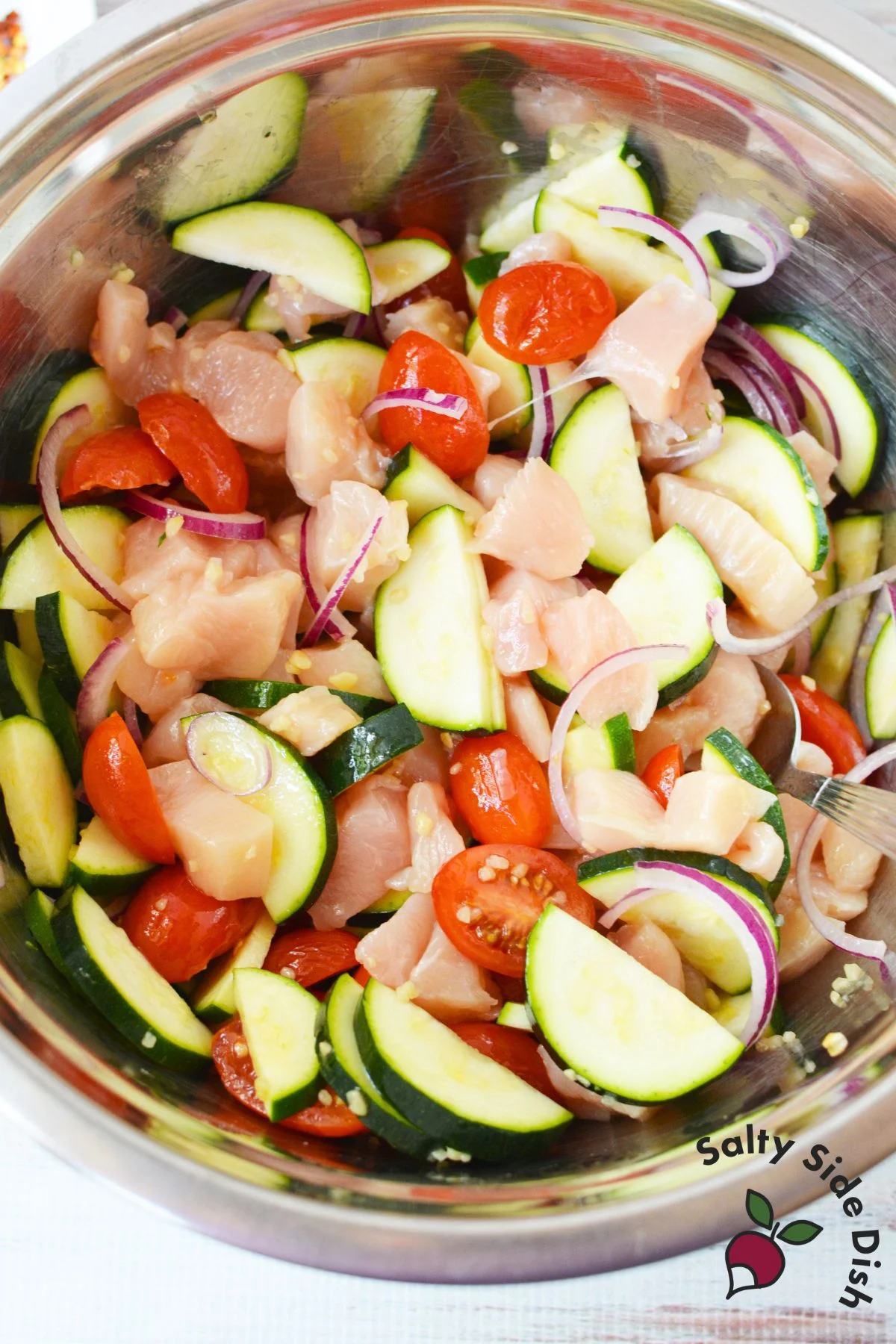 Bowl with raw chicken, zucchini, tomatoes, and onions mixed for Mediterranean chicken bake.