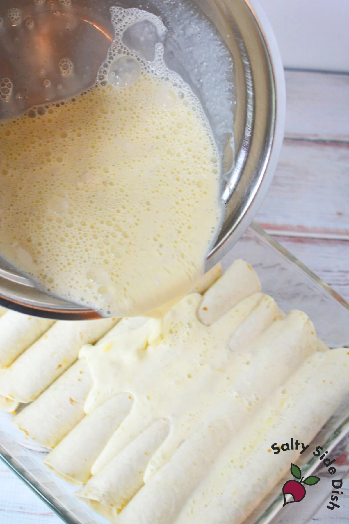 Whisked egg and cream mixture being poured over rolled tortillas in a baking dish to make a family breakfast casserole.
