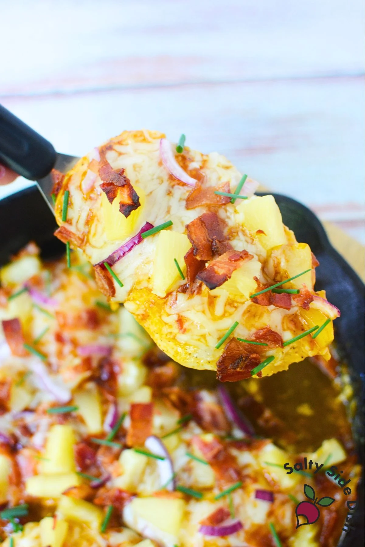 Serving a portion of baked Hawaiian chicken from a cast iron skillet with pineapple, bacon, and melted cheese.