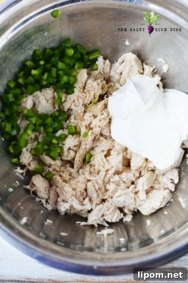 Cooked chicken mixed with diced jalapenos, mushrooms, sour cream, and shredded cheese in a bowl.