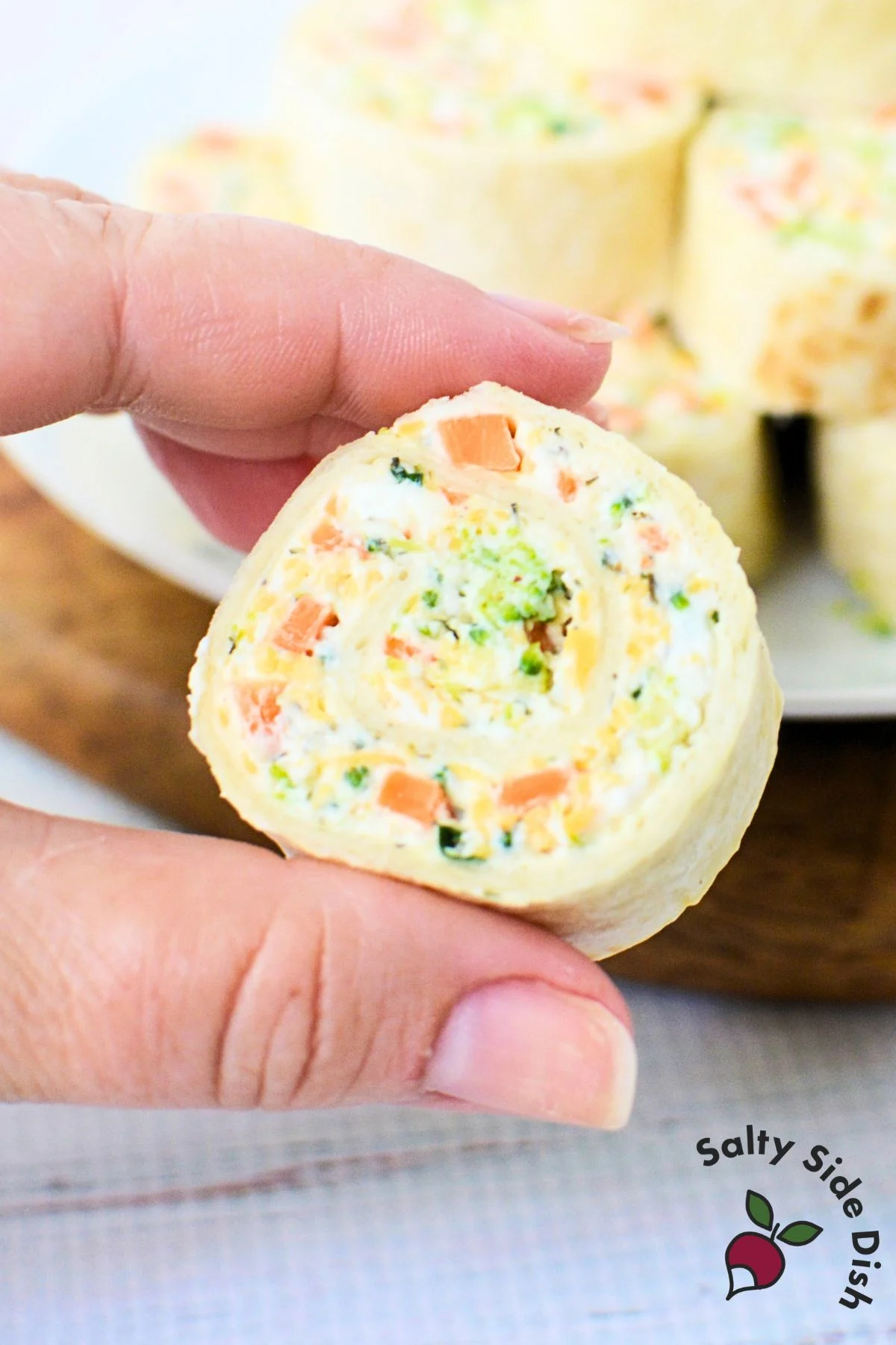 Hand holding a single veggie pinwheel with visible carrots, broccoli, and cheese, showing creamy texture and swirl of ingredients.