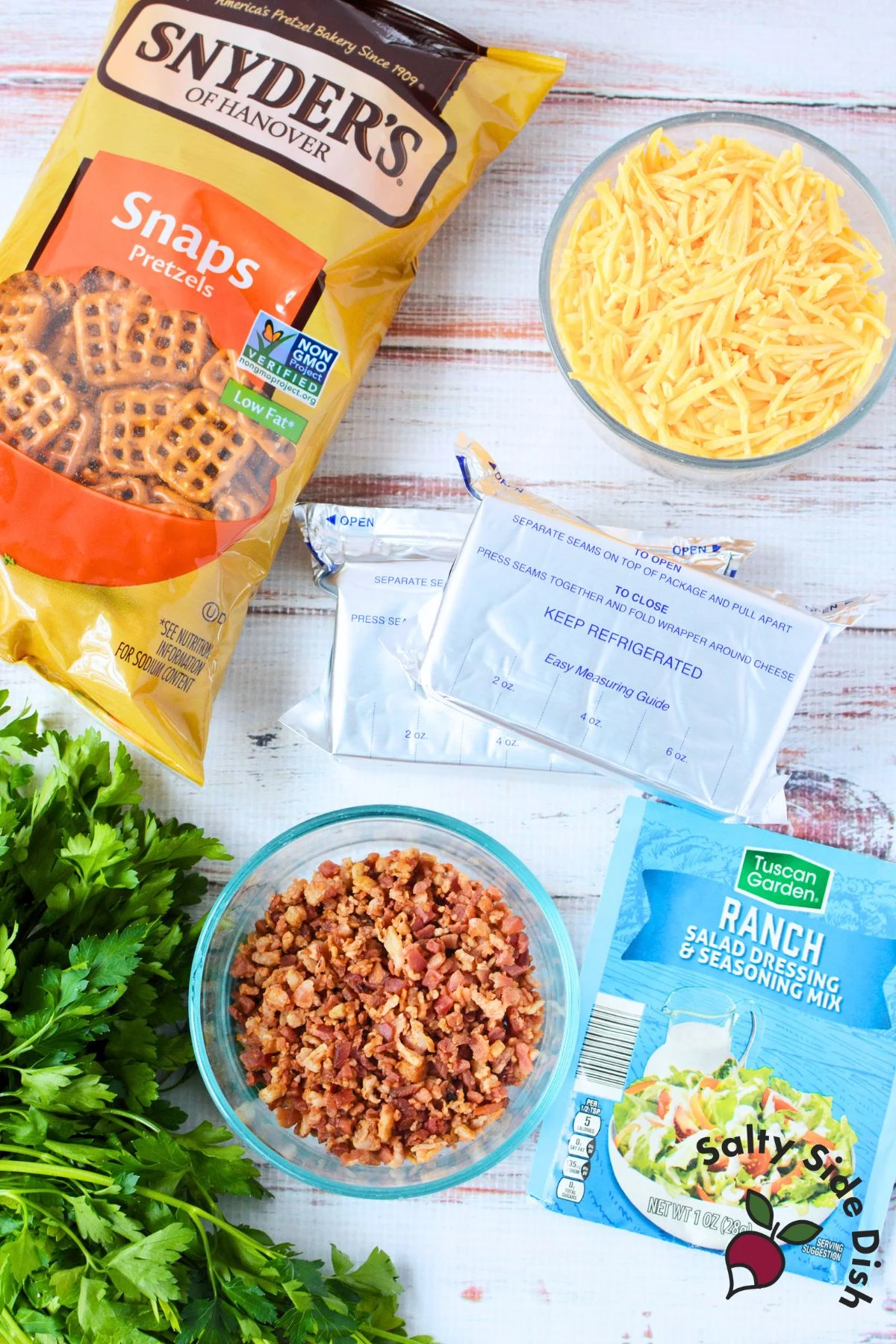 Ingredients for mini cheeseball bites including cream cheese, cheddar, ranch seasoning, bacon bits, parsley, and Snyder’s pretzel snaps.