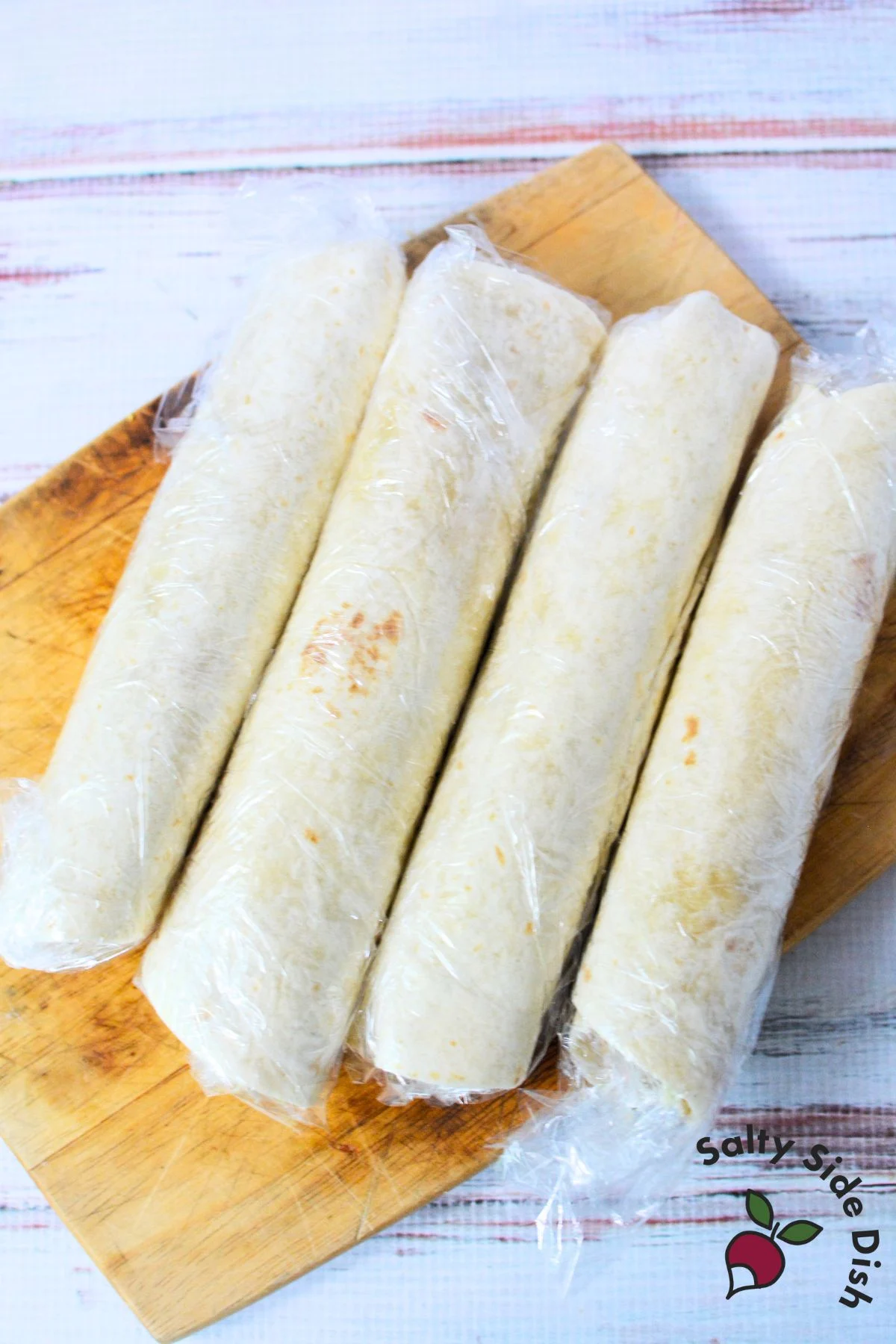 Tortilla pinwheel rolls wrapped tightly in plastic wrap and chilled on a wooden cutting board before slicing.