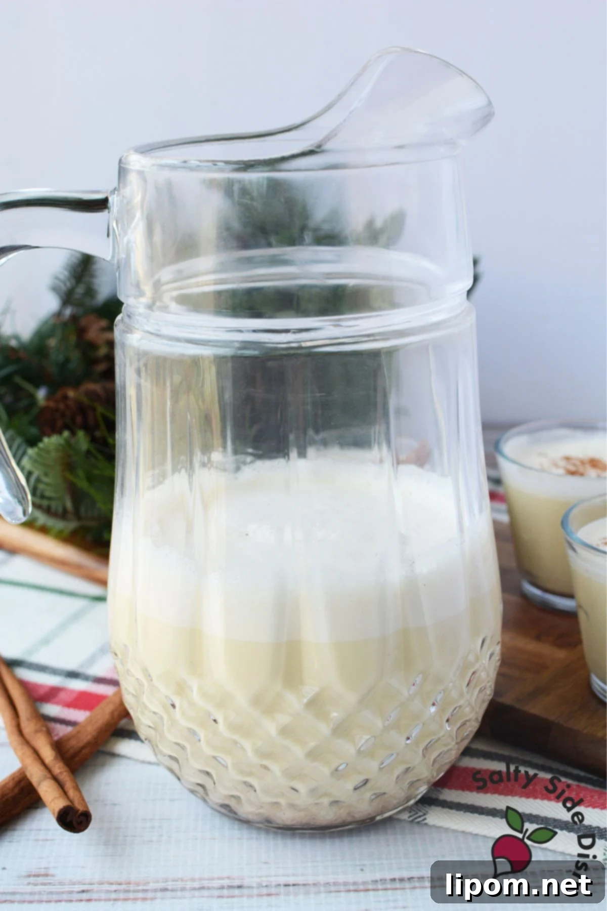 Spiked Holiday Cheer 5 A glass pitcher filled with homemade spiked eggnog gracing a holiday table, adorned with cinnamon sticks and lush Christmas greenery, creating a festive and inviting scene.