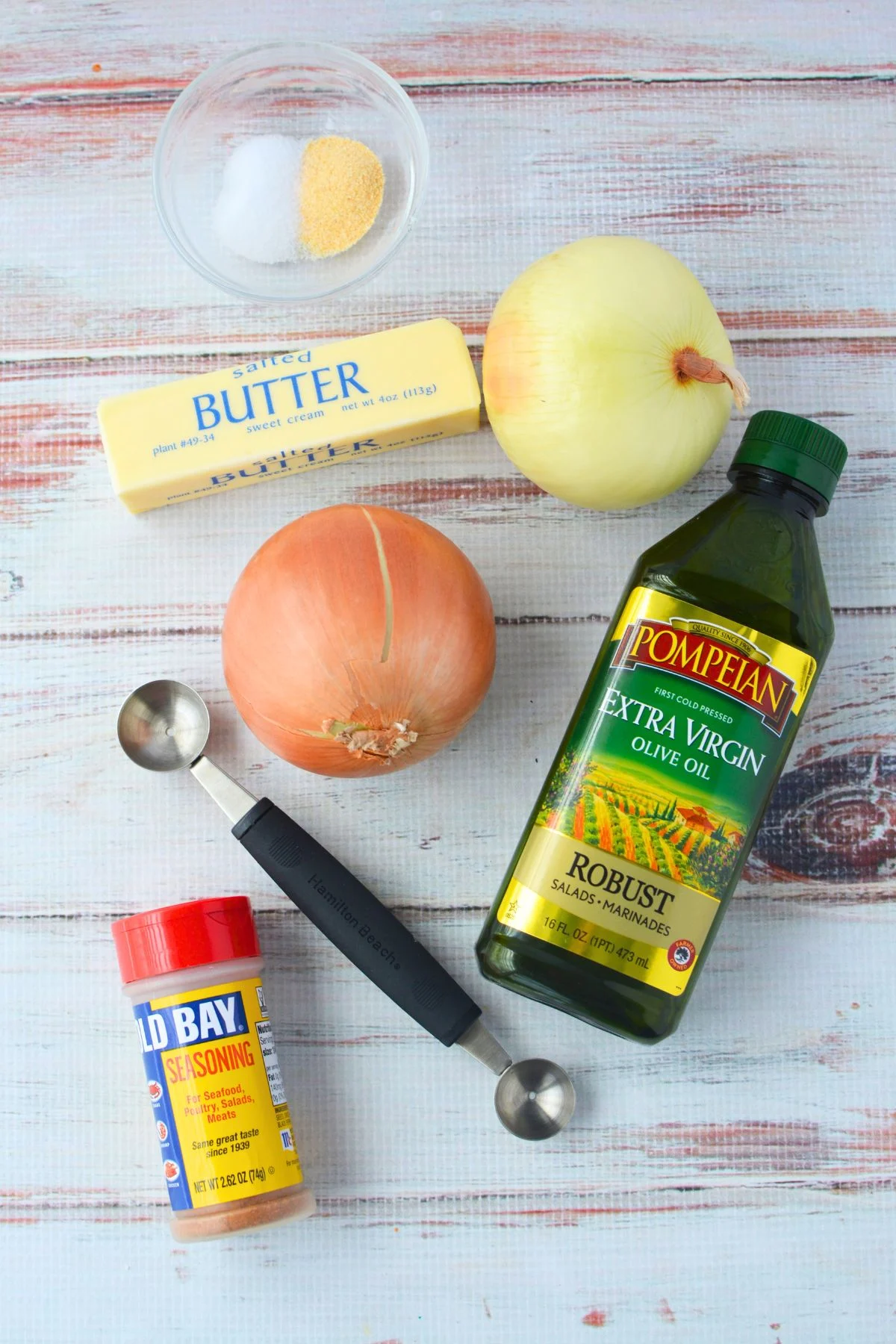Ingredients for onion boil recipe including sweet onions, butter, Old Bay seasoning, olive oil, garlic powder, salt, and a melon baller on a white wood surface