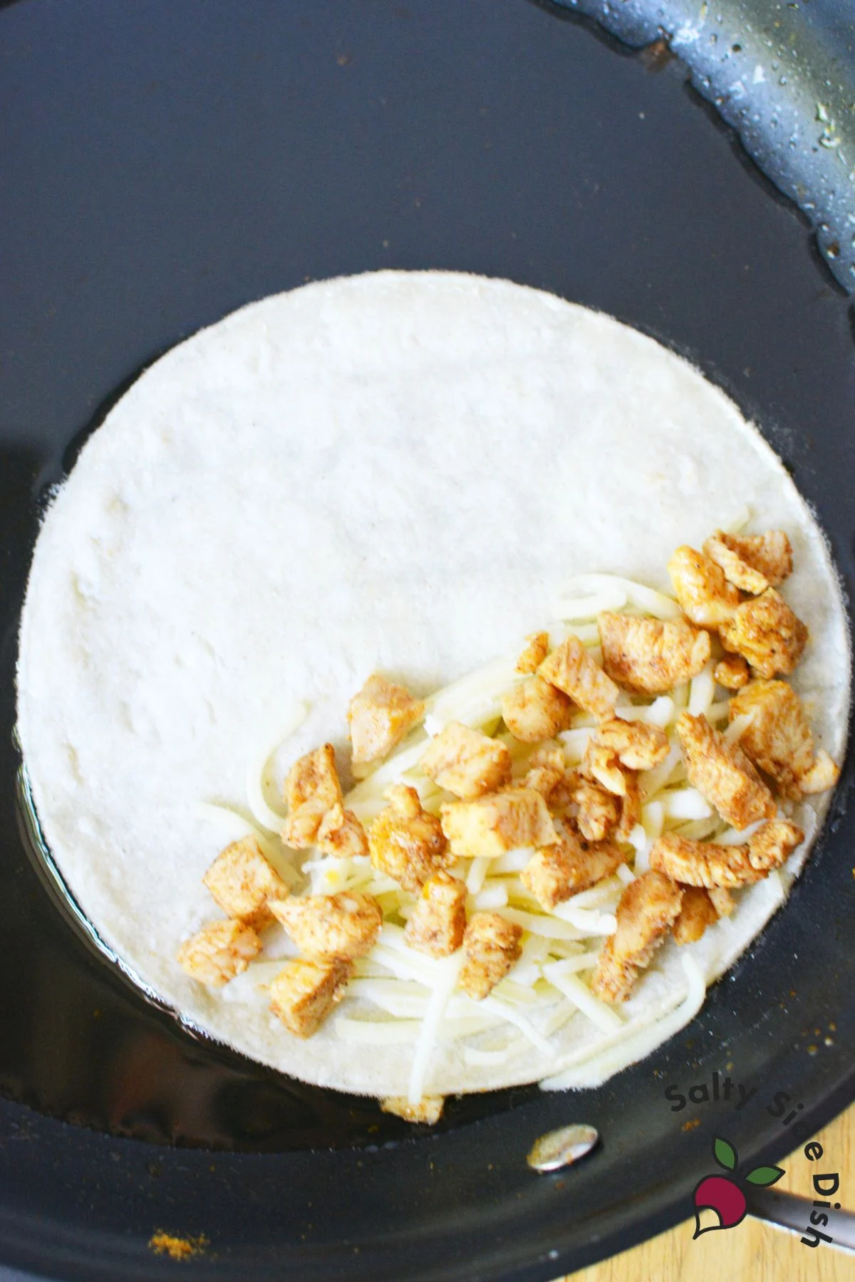 White corn tortilla in hot oil with shredded cheese and seasoned chicken on one half before folding