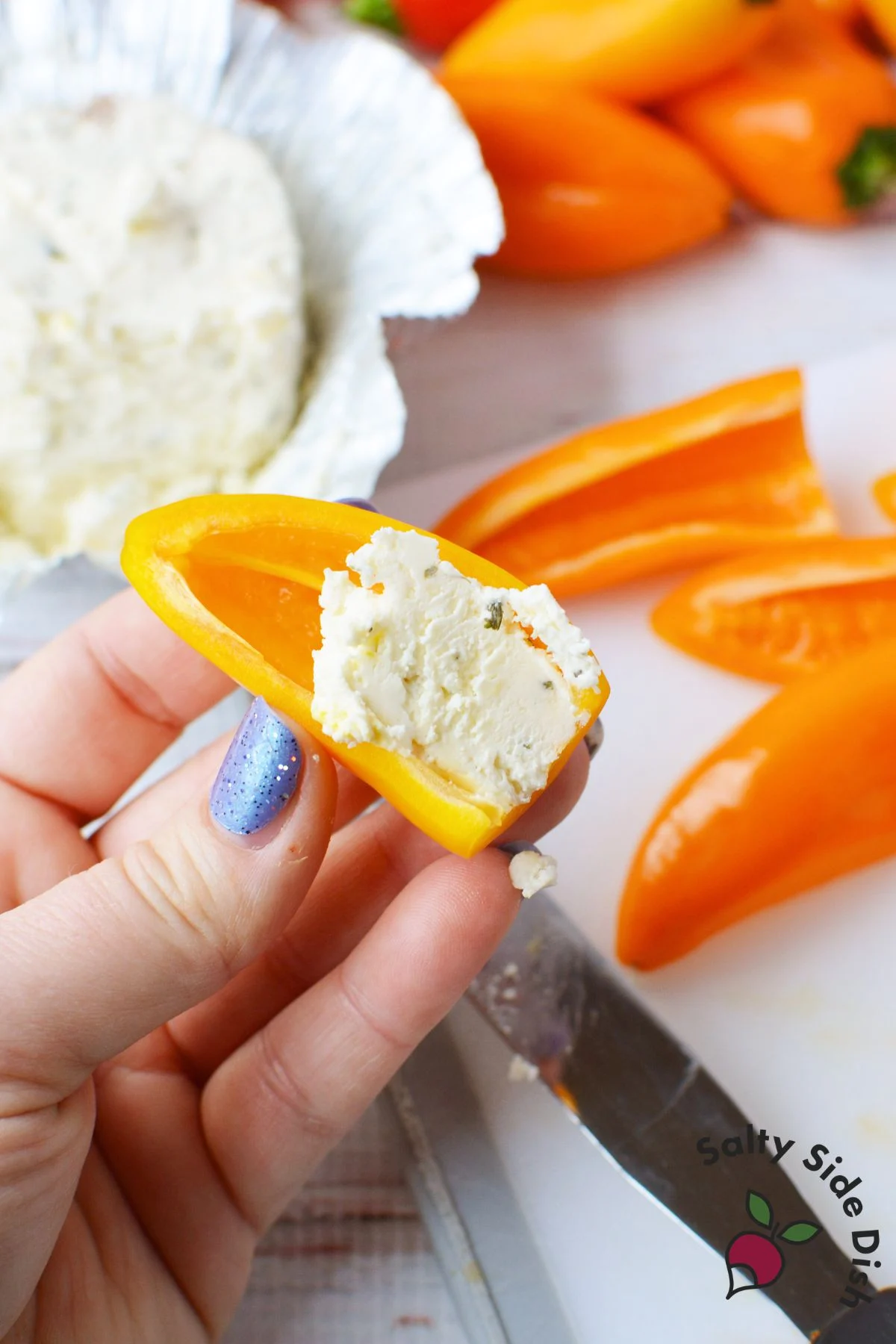 Hand pressing crumbly Boursin cheese into a yellow mini pepper half on a cutting board