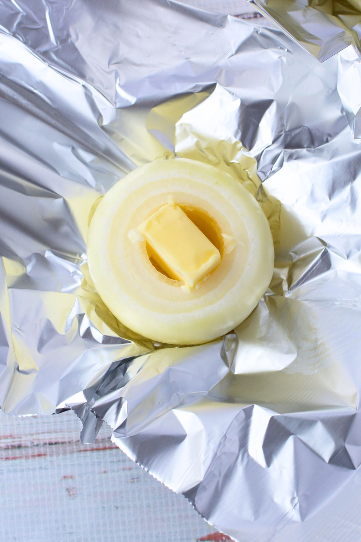 Two peeled sweet onions on a white cutting board, one hollowed out with a melon baller and one whole, with a stick of butter nearby