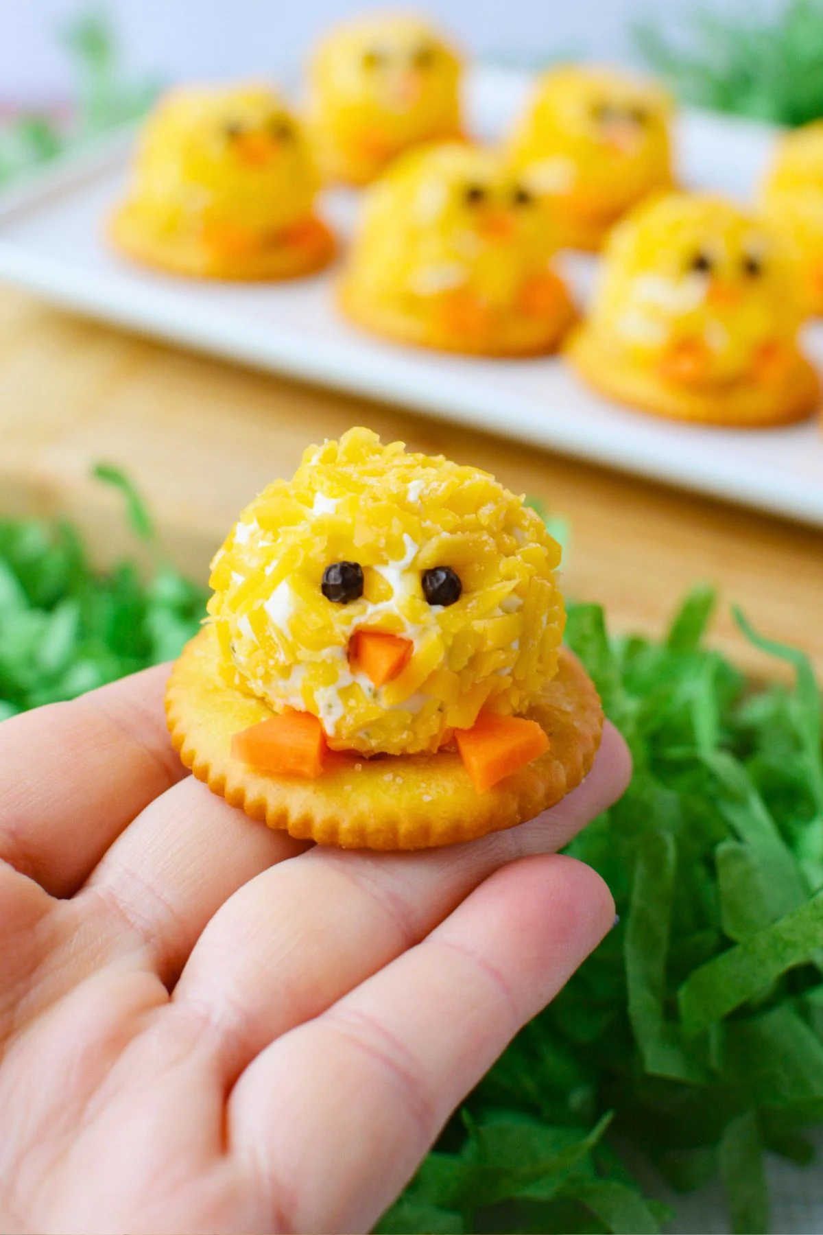 hand holding a mini chick cheeseball bite on a butter cracker with peppercorn eyes carrot beak and feet against a background of green Easter grass