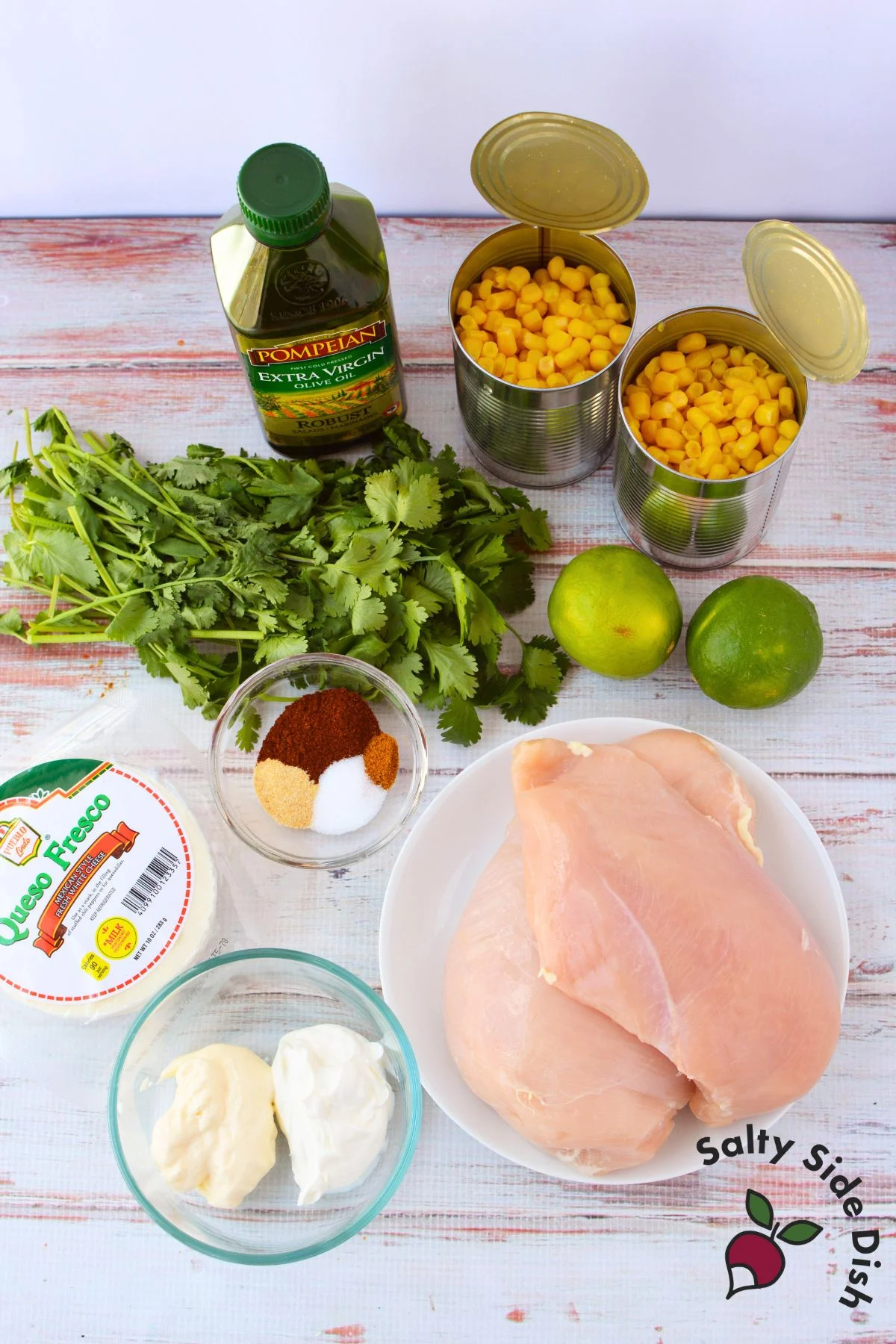 Ingredients for Mexican street corn chicken bake including canned corn, chicken breasts, queso fresco, olive oil, limes, cilantro, sour cream, mayonnaise, and spices
