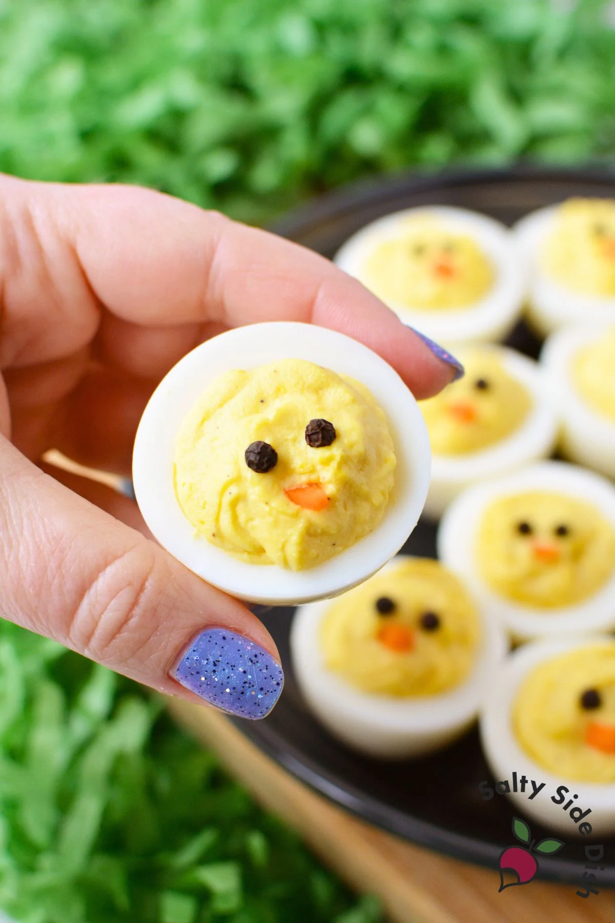 hand holding a single chick deviled egg with peppercorn eyes and carrot beak with a platter of Easter deviled eggs in the background