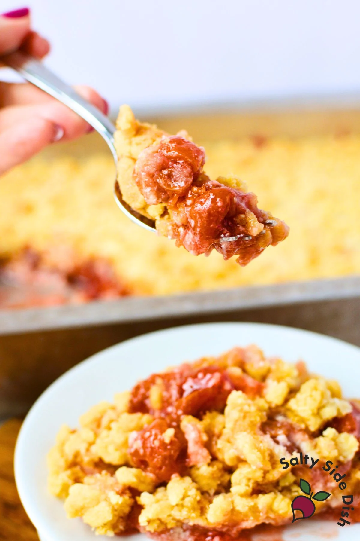 spoonful of cherry cheesecake dump cake held over a white plate with baking pan of golden crumble topping in background
