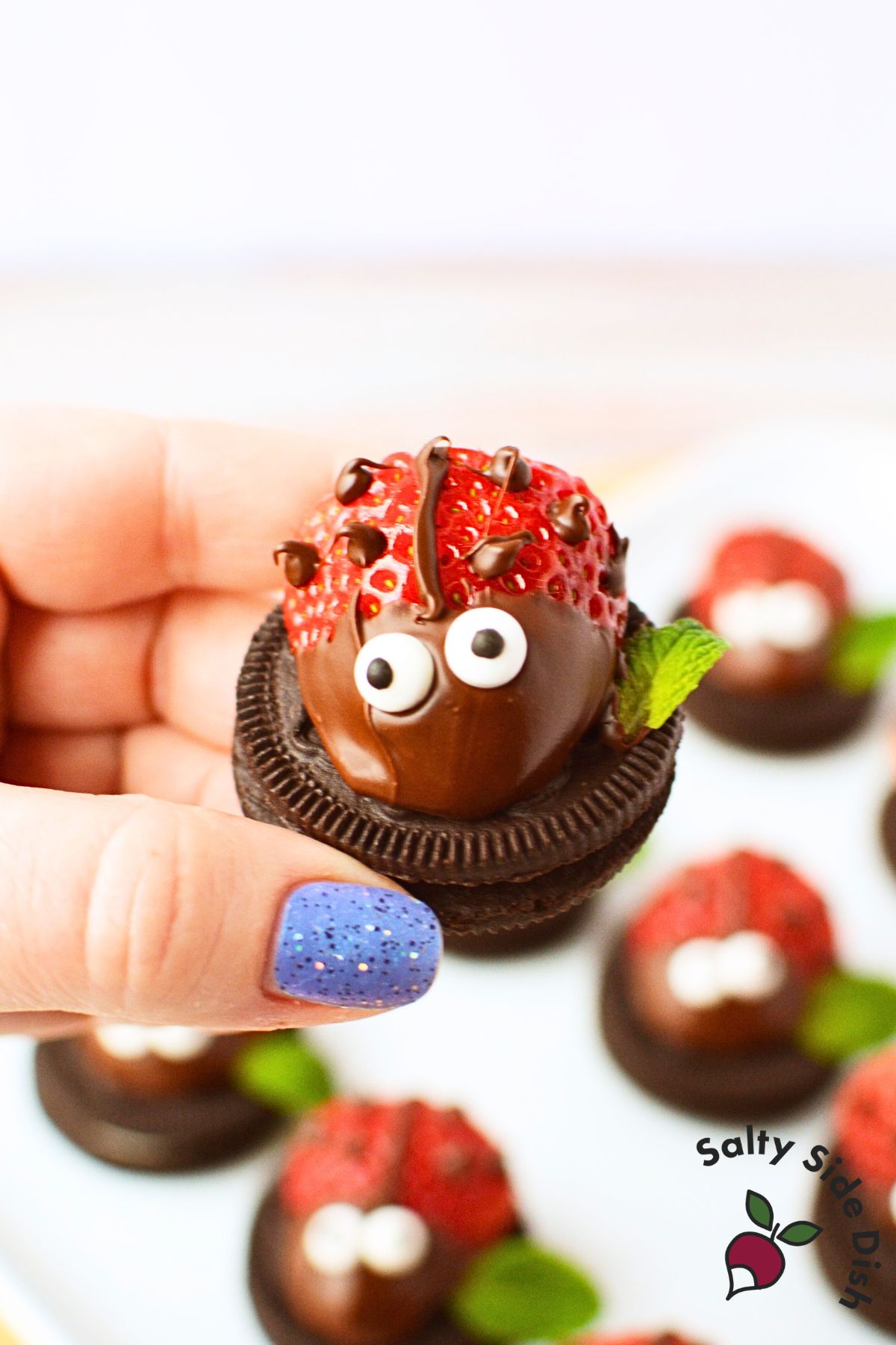 Hand holding a finished strawberry ladybug on an Oreo cookie base with chocolate chip spots, piped antennae, and a mint leaf wing