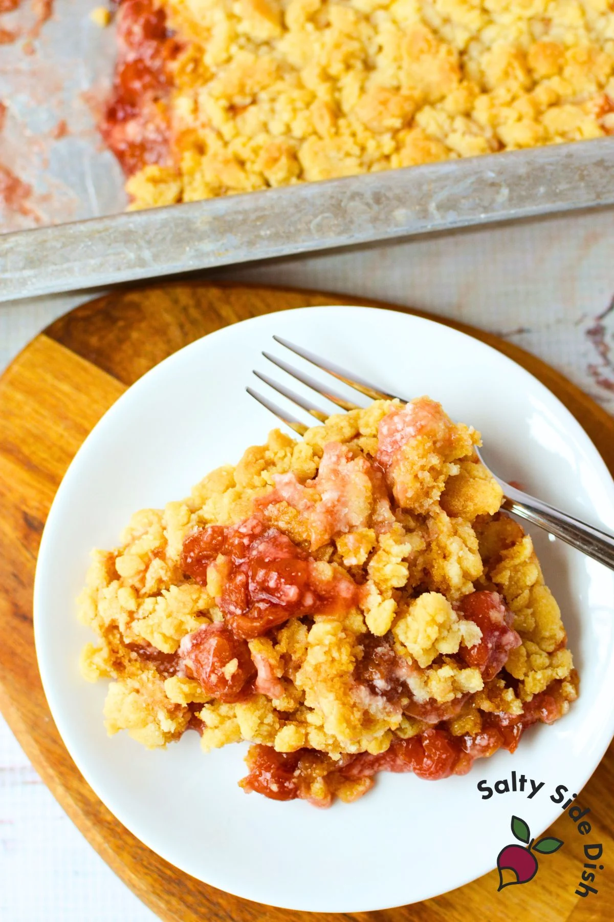 overhead view of cherry cheesecake dump cake serving on white plate with fork on wooden cutting board, baking pan in background