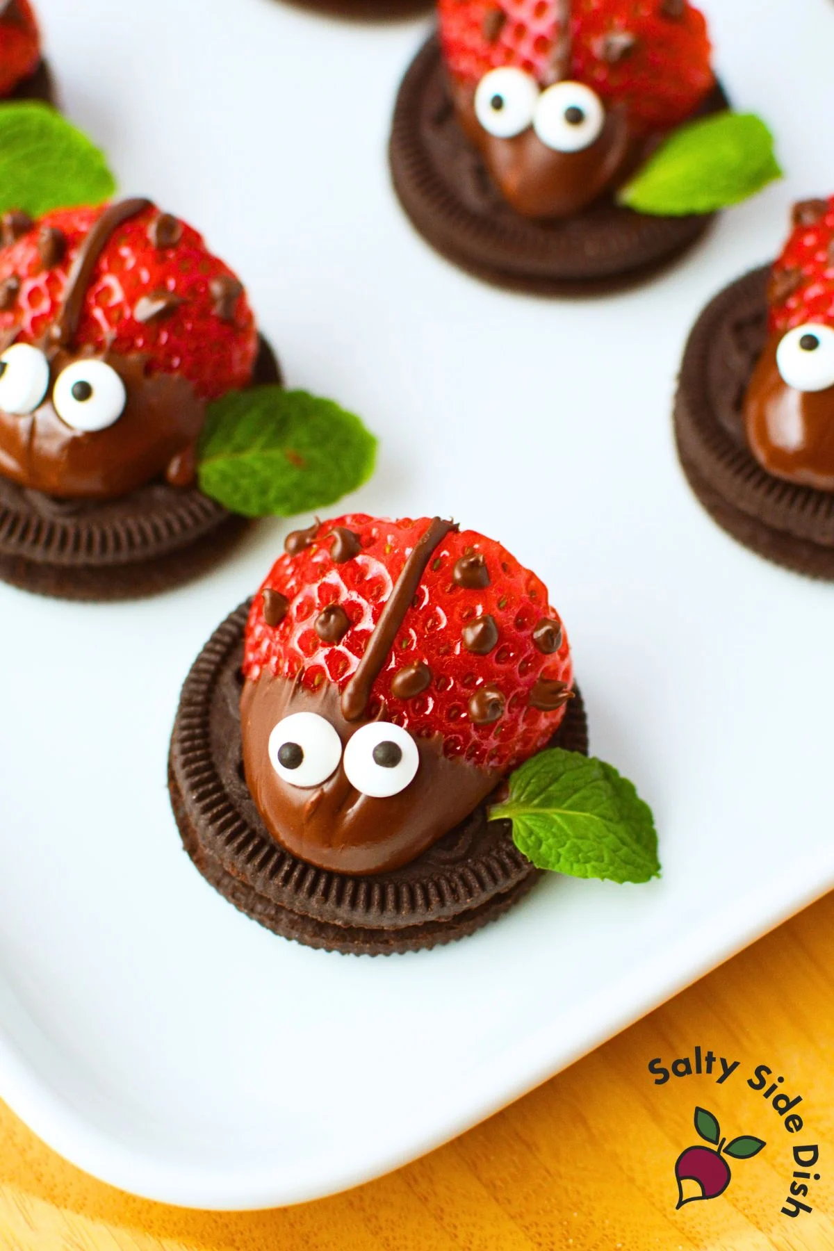 Single strawberry ladybug on an Oreo cookie with chocolate spots, candy eyes, and a fresh mint leaf on a white serving platter