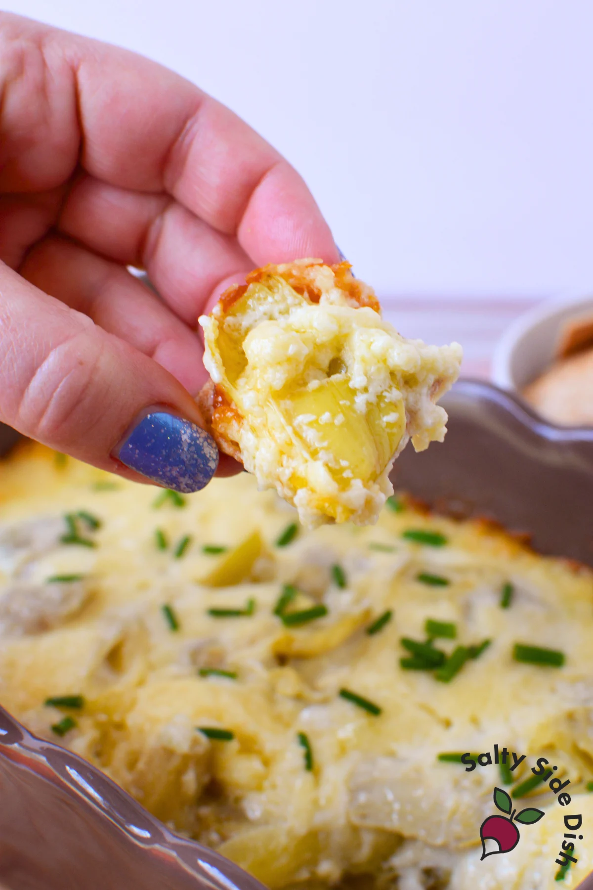 hand holding a cracker loaded with warm baked artichoke dip over a casserole dish