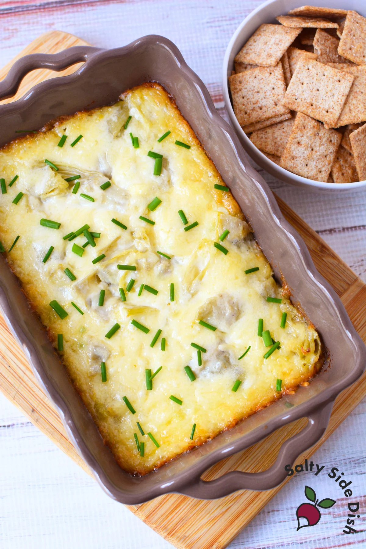 aked artichoke dip with golden bubbly top and fresh chives in a casserole dish served with wheat crackers