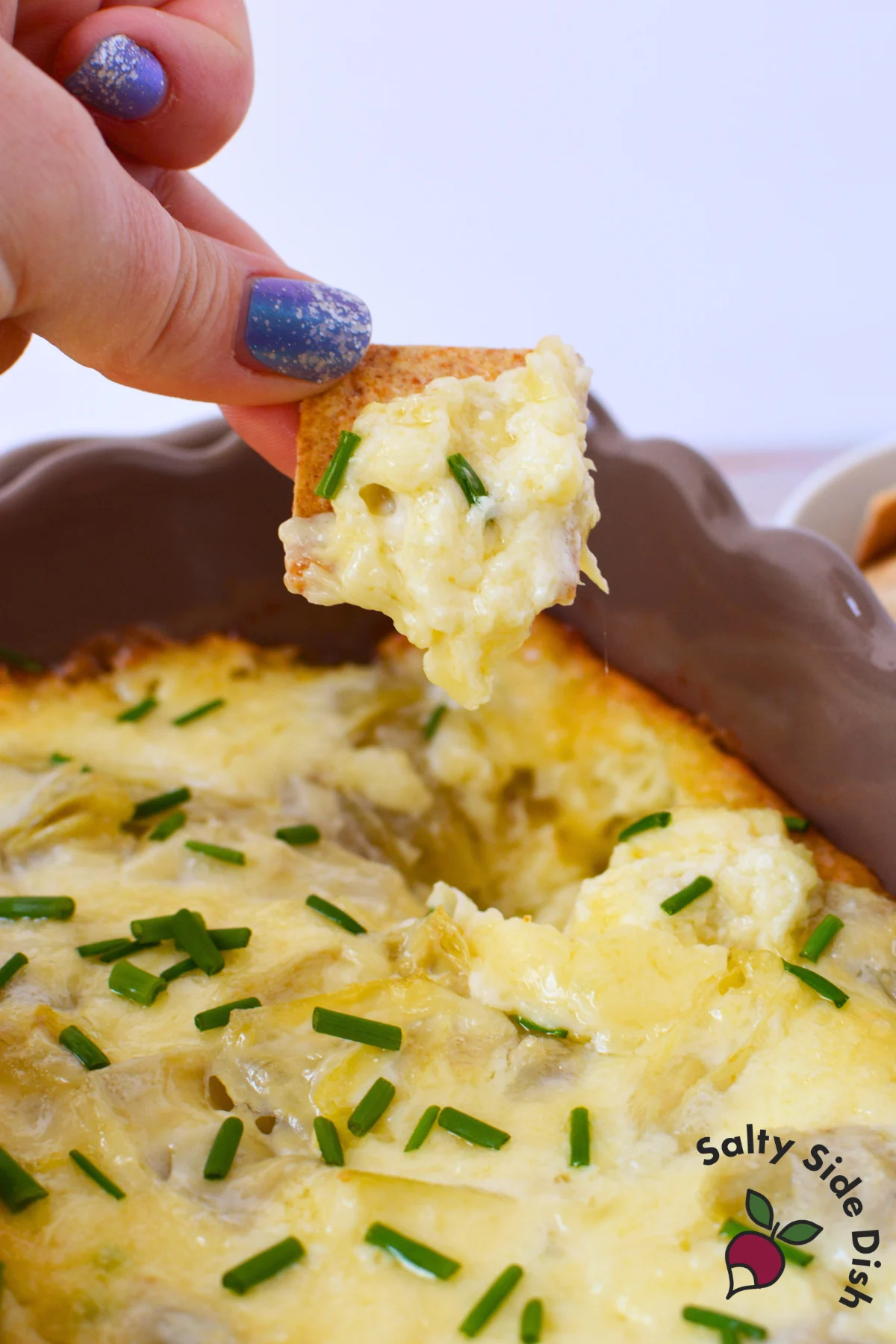 cracker being pulled from a round baking dish filled with hot bubbly artichoke dip topped with chives