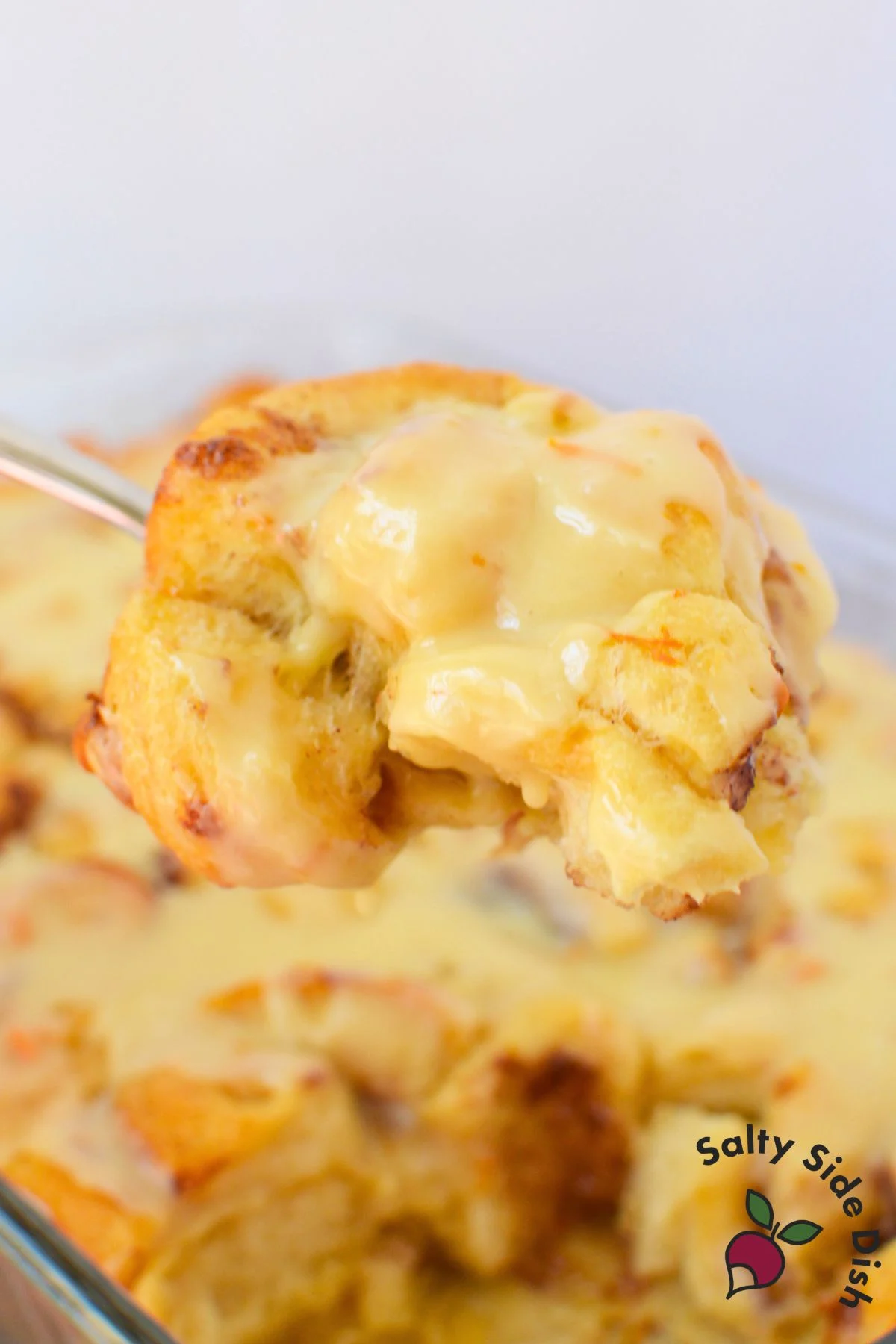 A large spoonful of orange roll casserole dripping with warm vanilla orange glaze held above the baking dish.