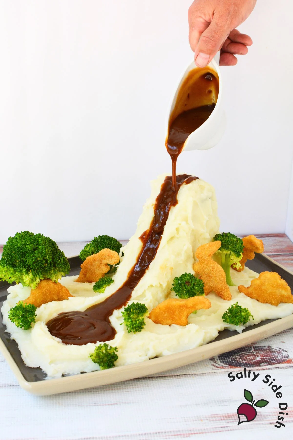 Hand pouring brown gravy down a mashed potato mountain surrounded by broccoli trees and dino nuggets