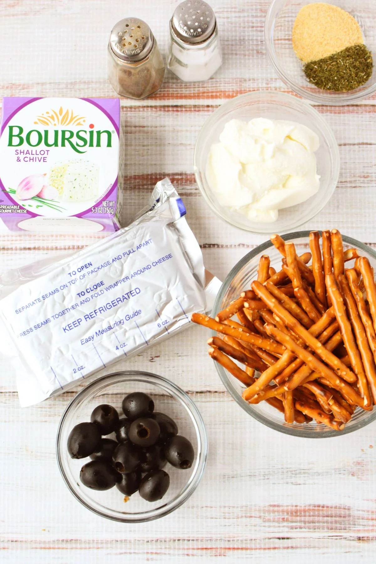 overhead view of hedgehog cheeseball ingredients including Boursin shallot and chive, cream cheese, black olives, pretzel sticks, and seasonings