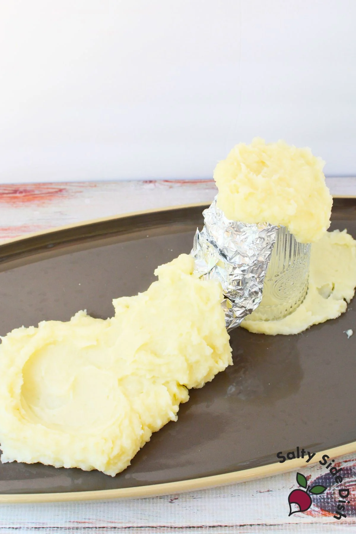 Mashed potatoes spread up the sides of a foil wrapped glass while building a potato volcano