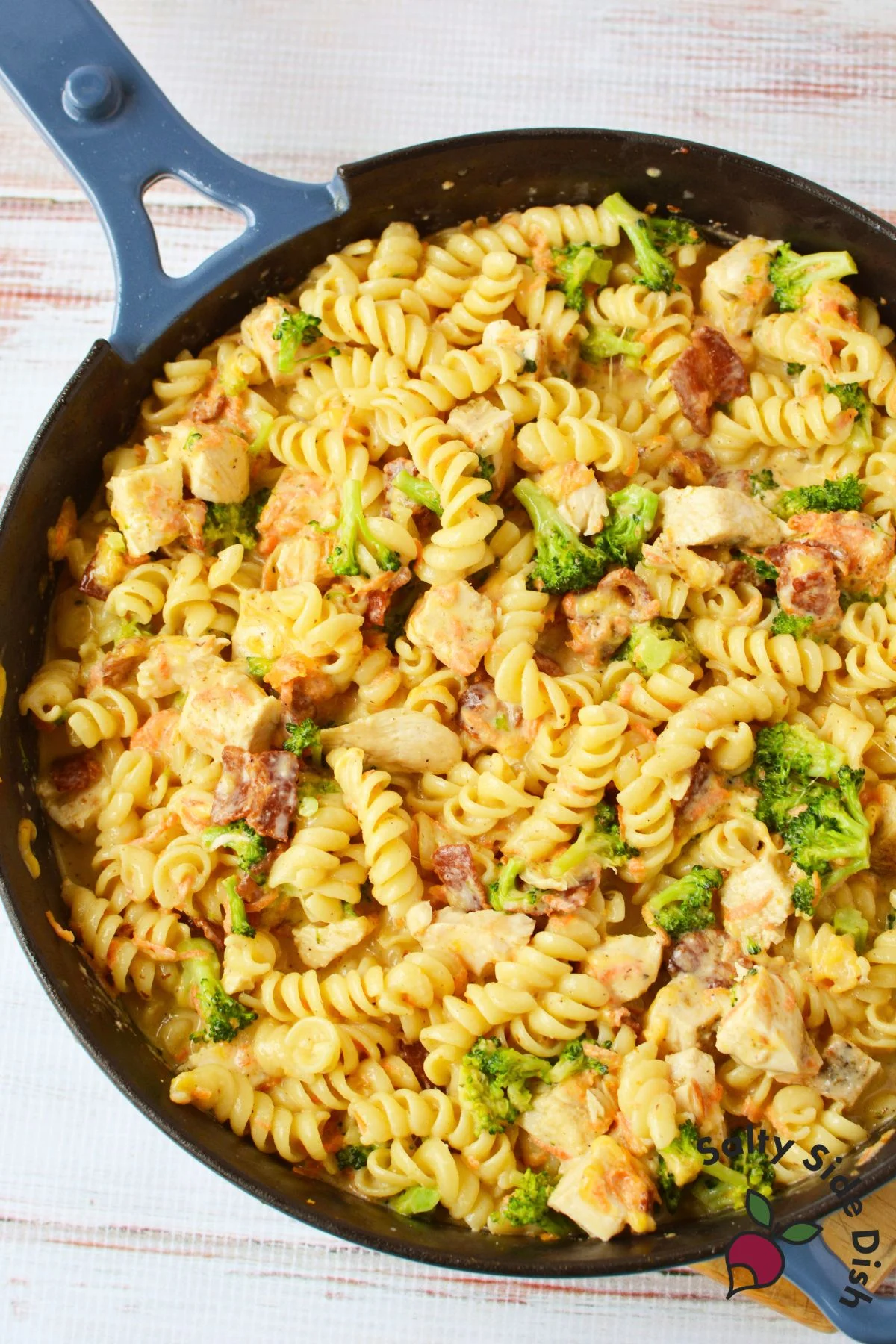 Skillet filled with creamy rotini pasta, chicken, broccoli, bacon, carrots, and melted cheese.