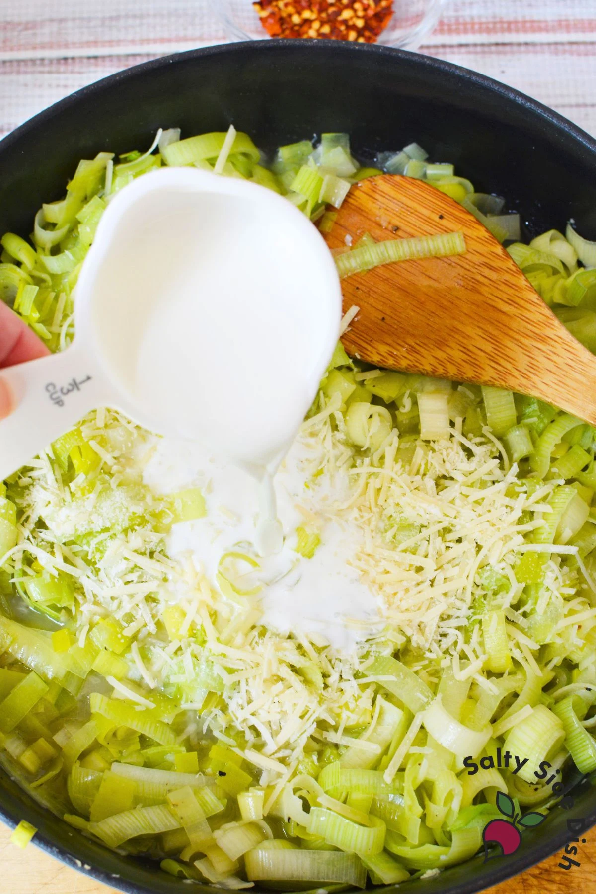 Pouring half and half over cooked leeks topped with shredded parmesan in a skillet